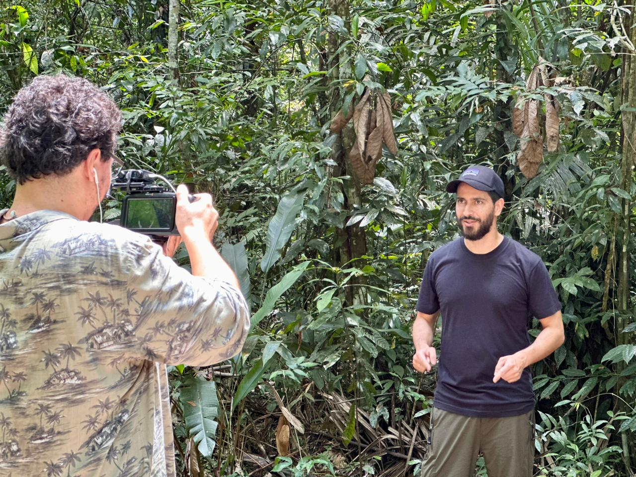 Um fotógrafo segura a câmera na direção de outro homem, que está de bonés e camisa pretos. Ao fundo, a vegetação em verde escuro.