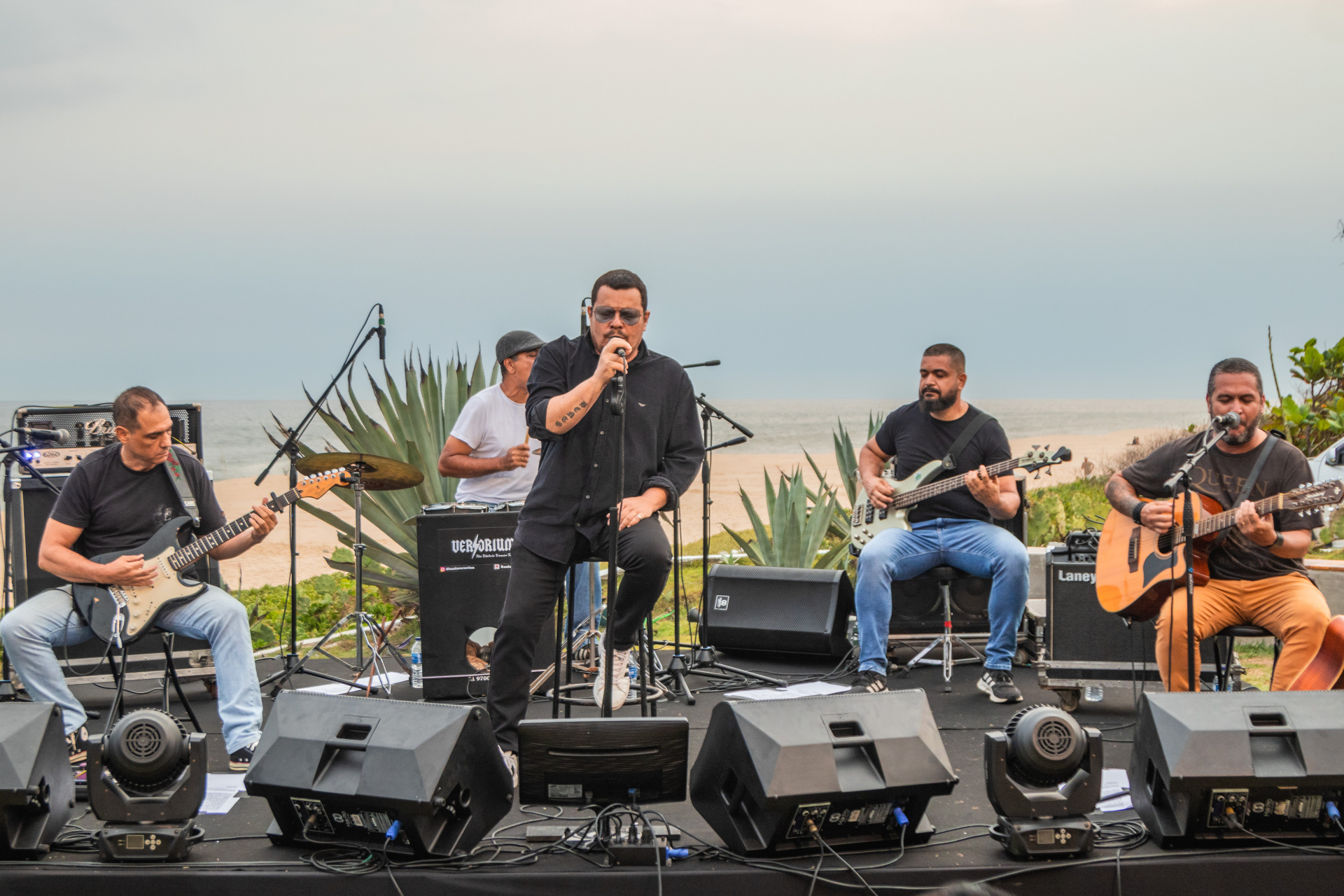 Cinco musicistas estão sentados, se apresentando. Ao fundo, o mar.