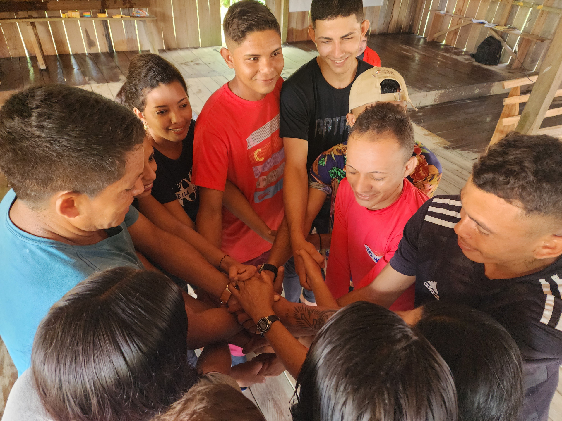 Jovens unem suas mãos no centro do círculo que formam. Todos sorriem.