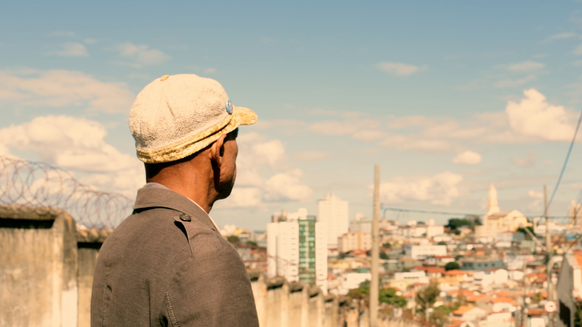 Um homem é visto por trás. Ele usa boné e olha para os prédios de uma cidade, ao fundo. O céu está azul, com algumas nuvens.