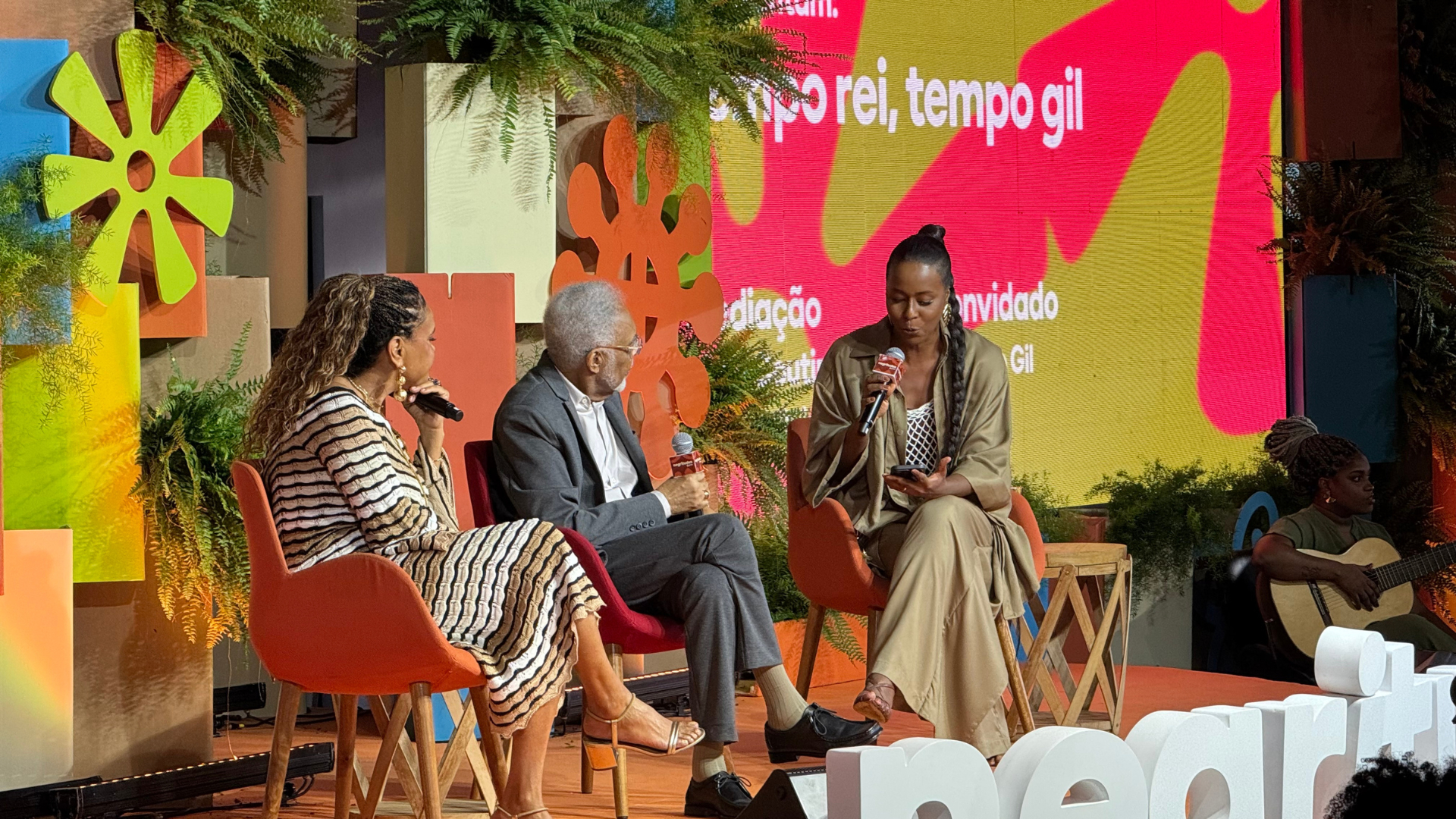 Gilberto Gil é entrevistado por Maju Coutinho e Teresa Cristina durante o painel “Tempo Rei, Tempo Gil”, no palco do Negritudes Globo. Os três estão sentados em cadeiras laranjas, com microfones nas mãos, diante de um cenário vibrante com plantas e formas coloridas. Ao fundo, uma telona exibe o nome do painel e os nomes dos convidados.