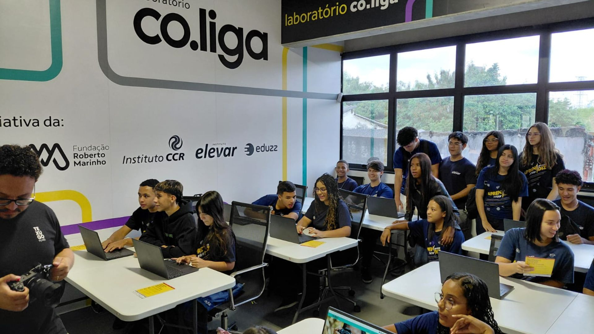 Turma de jovens participa de atividade no laboratório co.liga, em um ambiente moderno e colaborativo. Sentados em mesas com notebooks, os alunos interagem entre si com entusiasmo, enquanto outros observam em pé, formando um grupo diverso e engajado. Ao fundo, uma parede destaca as marcas apoiadoras do projeto, como Fundação Roberto Marinho, Instituto CCR, Instituto Elevar e Eduzz. A atmosfera é de aprendizado dinâmico, criatividade e inclusão, com foco na cultura digital e tecnologia.