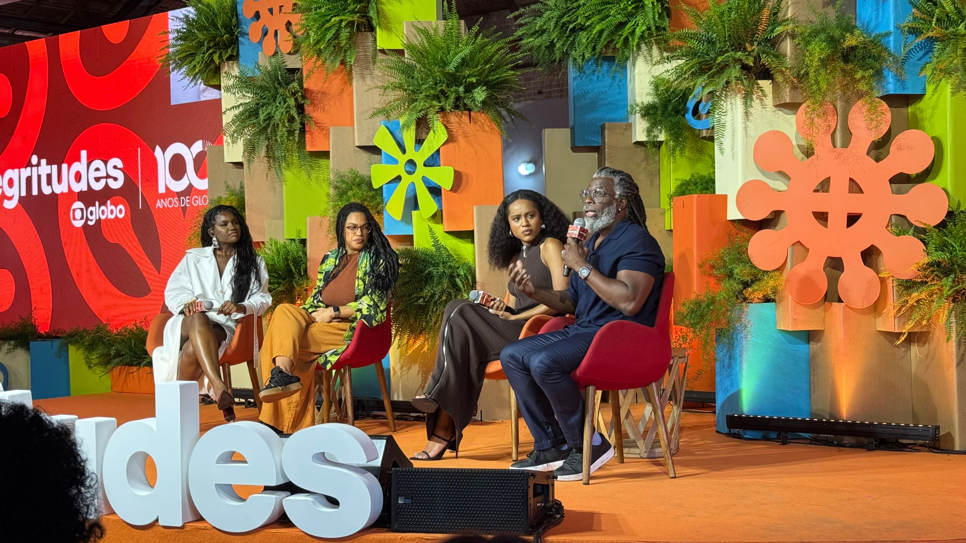 Quatro pessoas negras participam de um painel no palco do Negritudes Globo. Entre elas estão duas mulheres e dois homens, sentados em cadeiras coloridas, com microfones nas mãos. Um dos participantes fala enquanto os outros ouvem atentamente. O cenário é colorido, decorado com samambaias e painéis geométricos. Ao fundo, o telão mostra a marca do evento e os “100 anos de Globo”.