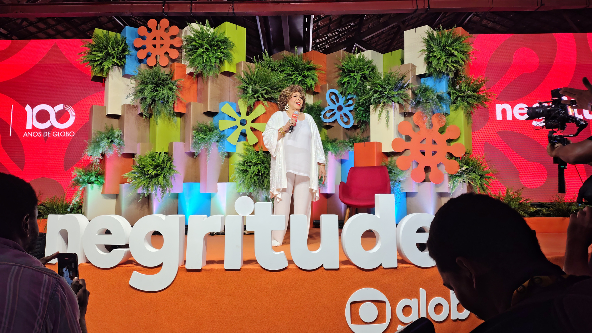 A cantora Alcione está no centro do palco do Negritudes Globo, sorrindo e segurando um microfone. Ela veste um conjunto branco elegante e está cercada por uma decoração colorida com plantas, flores estilizadas e um letreiro grande escrito "negritudes" à sua frente. O fundo do palco exibe a identidade visual do evento e a marca “100 anos de Globo”