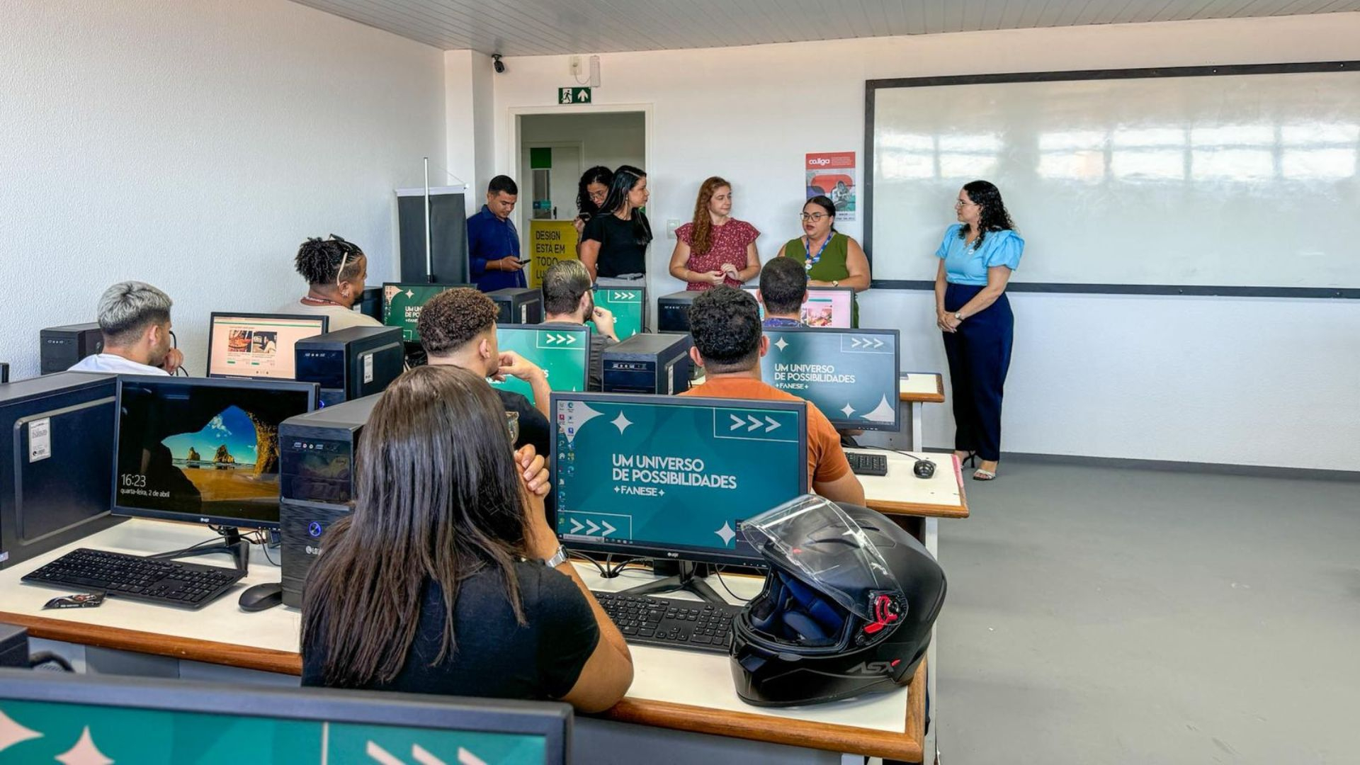 Sala de aula de informática com vários alunos sentados em frente a computadores de mesa. Na parte frontal da sala, cinco pessoas — possivelmente professoras ou palestrantes — estão em pé, conversando com os estudantes. As telas dos computadores exibem a frase “UM UNIVERSO DE POSSIBILIDADES + FANESE”, indicando que o local pertence à instituição de ensino FANESE. O ambiente é iluminado, moderno e organizado, com um quadro branco grande ao fundo. Um capacete de motociclista está sobre uma das mesas, sugerindo