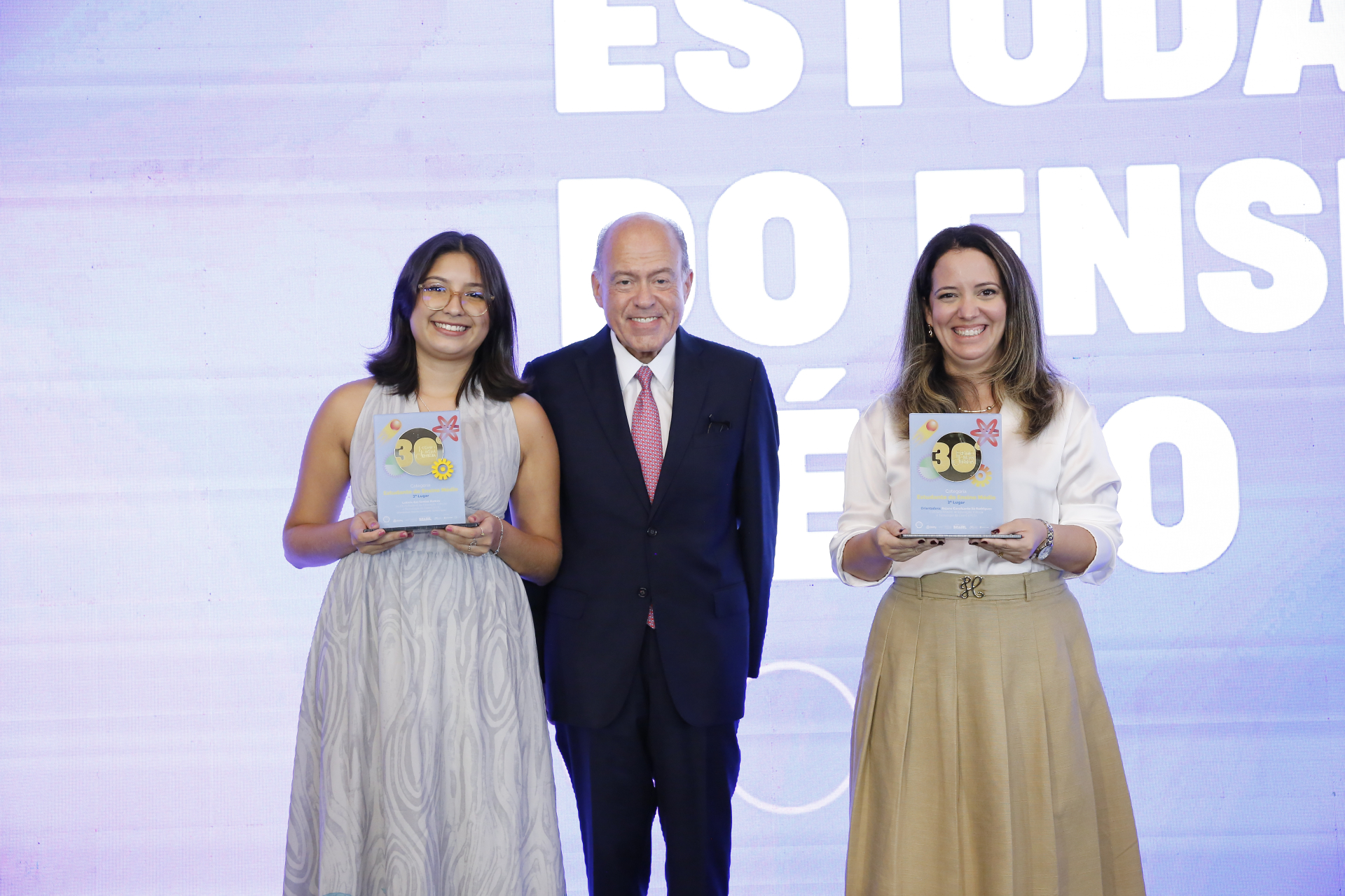 Na imagem, três pessoas posam sorridentes para a foto durante uma cerimônia de premiação. Ao centro, está um homem mais velho, vestindo terno azul-marinho e gravata rosa. À esquerda, uma jovem mulher de cabelo liso na altura dos ombros, vestindo um vestido cinza claro e segurando um troféu. À direita, uma mulher adulta de cabelos longos, vestindo blusa branca e saia bege, também segurando um troféu. Ao fundo, um painel iluminado exibe parte da frase “ESTUDANTES DO ENSINO MÉDIO”