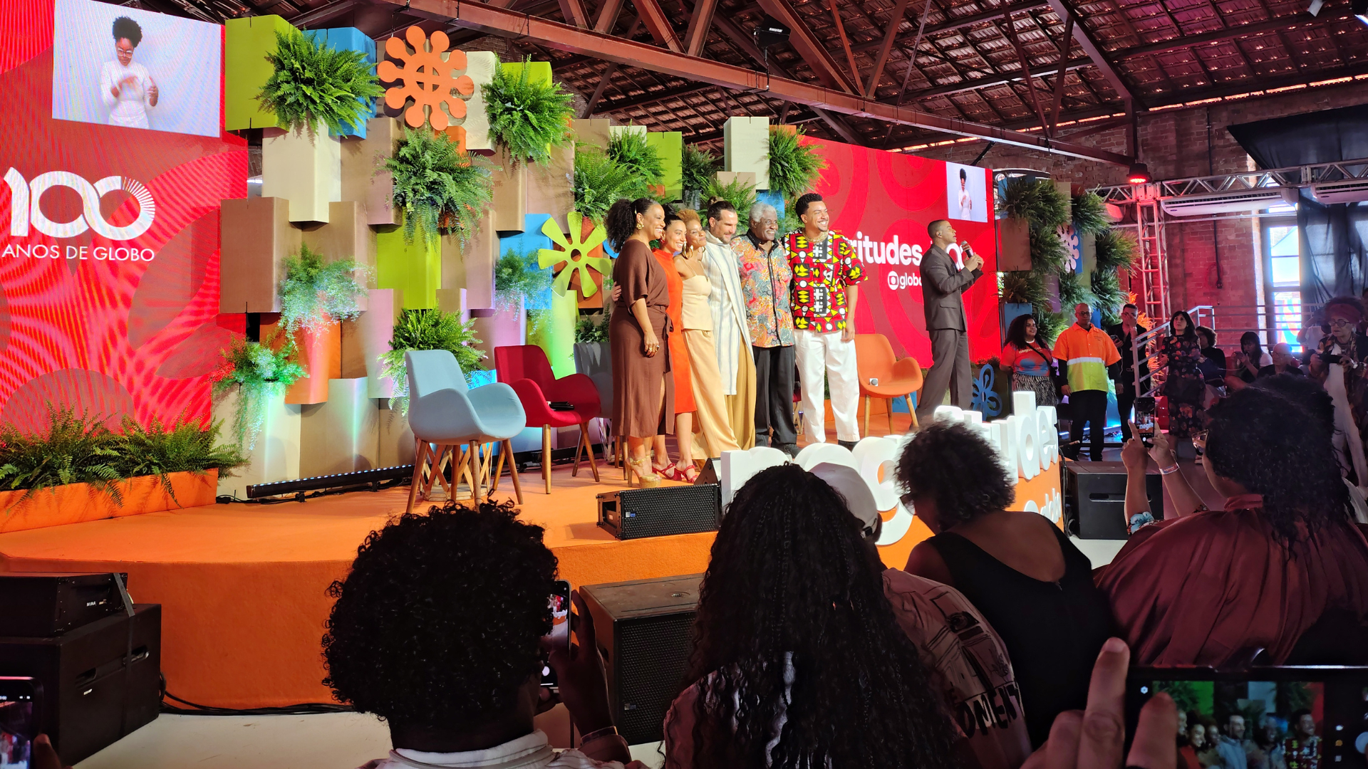 Grupo de sete pessoas posa sorridente no palco do evento Negritudes Globo, diante de um cenário decorado com plantas e painéis coloridos. As pessoas estão abraçadas, lado a lado, em clima de celebração. À direita, um homem fala ao microfone. A plateia acompanha e registra o momento com celulares. No fundo, aparece o logotipo "100 anos de Globo" e a identidade visual do evento.