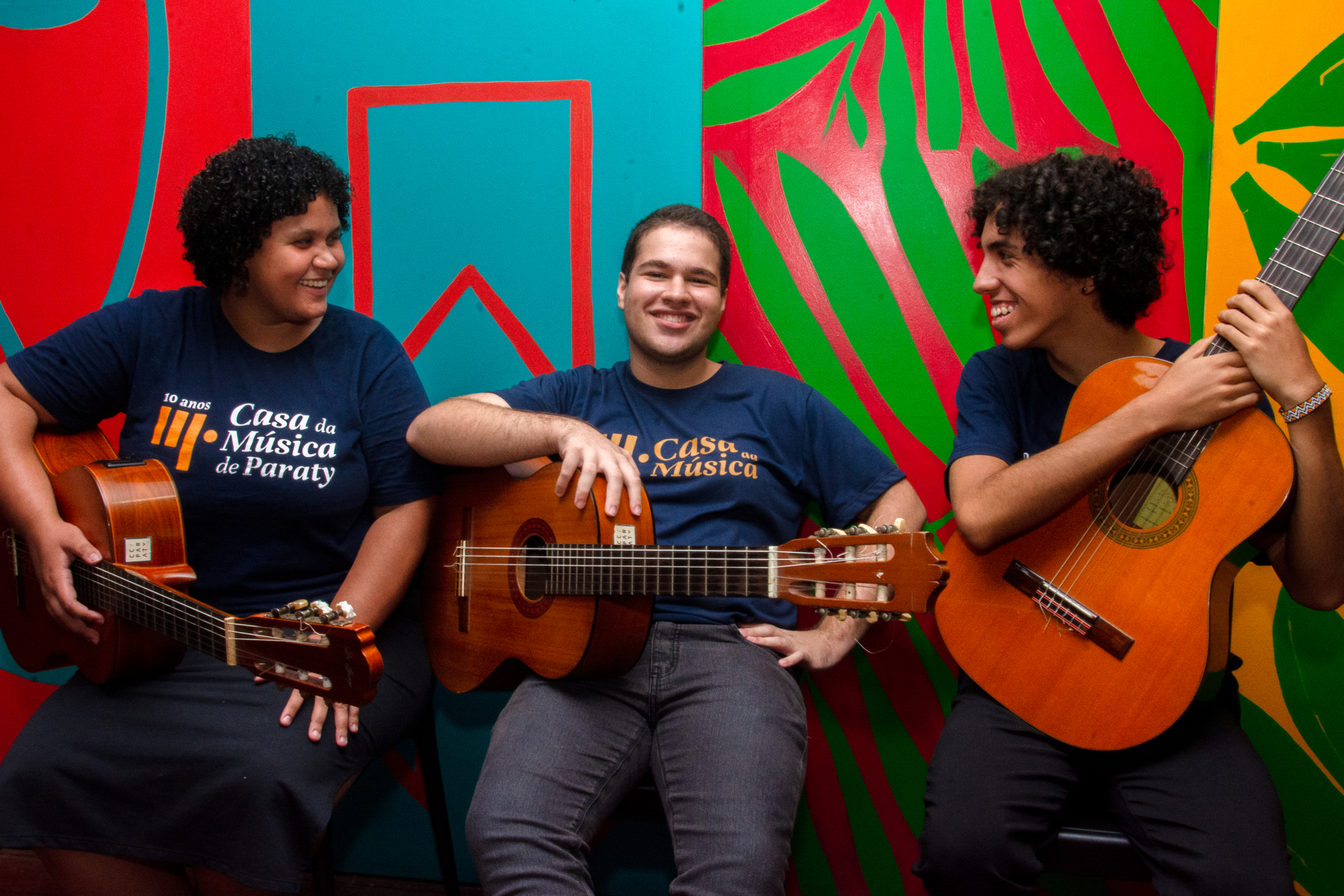 Três jovens músicos estão sentados lado a lado em frente a uma parede colorida com formas abstratas em azul, vermelho, verde e amarelo. Todos sorriem e seguram violões. À esquerda, uma jovem de cabelo cacheado curto veste uma camiseta azul-marinho com o logo dos “10 anos da Casa da Música de Paraty”. No centro, um rapaz também veste a camiseta do projeto e descansa o braço sobre o violão, sorrindo diretamente para a câmera. À direita, outro jovem de cabelo cacheado sorri olhando para o colega ao centro, se