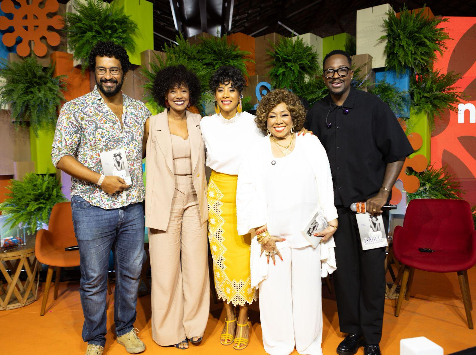 Cinco pessoas negras — três mulheres e dois homens — posam juntas sorrindo no palco do Negritudes Globo. Entre elas estão Alcione (ao centro, de branco), a jornalista Aline Midlej, a atriz Isabel Fillardis, Pedro Rajão e o músico Mumuzinho. Todos seguram um exemplar do livro "Isabel" nas mãos. O cenário é colorido, com folhagens e painéis vibrantes.