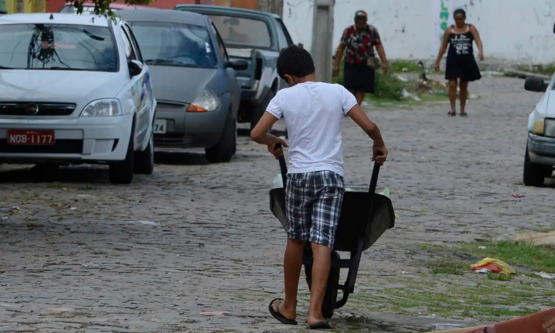 Um menino veste camiseta branca, bermuda xadrez e chinelos enquanto empurra um carrinho de mão vazio em uma rua de paralelepípedos. Ele está de costas e caminha entre carros estacionados. Ao fundo, há três mulheres andando na calçada. A cena sugere trabalho infantil em um ambiente urbano e simples.