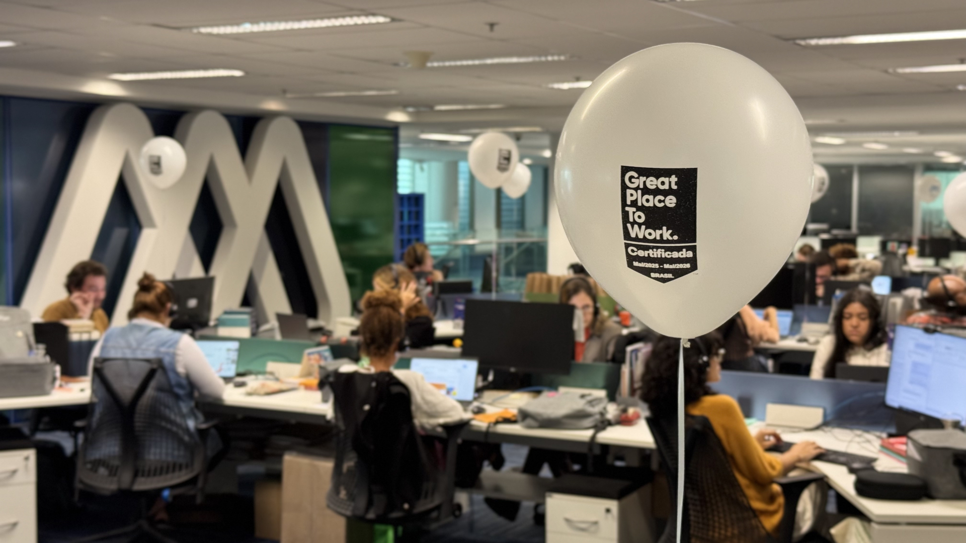 Ambiente de escritório moderno e colaborativo com diversas pessoas trabalhando em frente a computadores. Em destaque, há um balão branco com a inscrição "Great Place To Work – Certificada – Mai/2023 - Mai/2025 – BRASIL", celebrando a certificação da empresa como um excelente local para se trabalhar. Ao fundo, o espaço apresenta divisórias de vidro, móveis ergonômicos e um grande logo em branco estilizado na parede. O clima é dinâmico, produtivo e de celebração.