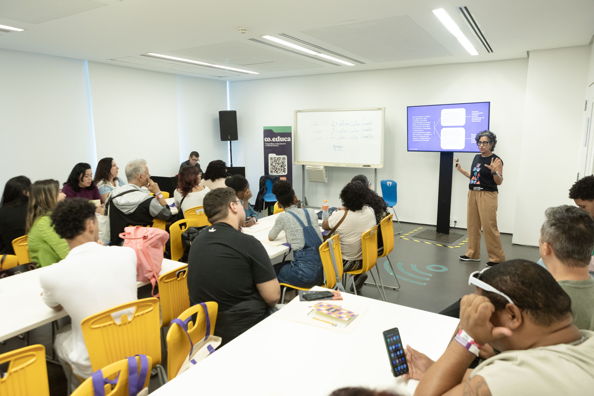 Em uma sala de aula bem iluminada, com paredes brancas e cadeiras amarelas, uma mulher de cabelos curtos e grisalhos, vestindo camiseta preta e calça bege, conduz uma apresentação em pé, gesticulando enquanto fala. À sua esquerda, uma televisão exibe um slide com fundo roxo e textos em branco. Os participantes, cerca de 20 pessoas, estão sentados em grupos em torno de mesas brancas, atentos ou fazendo anotações. Ao fundo, um quadro branco contém uma lista escrita à mão. O ambiente transmite concentração e e