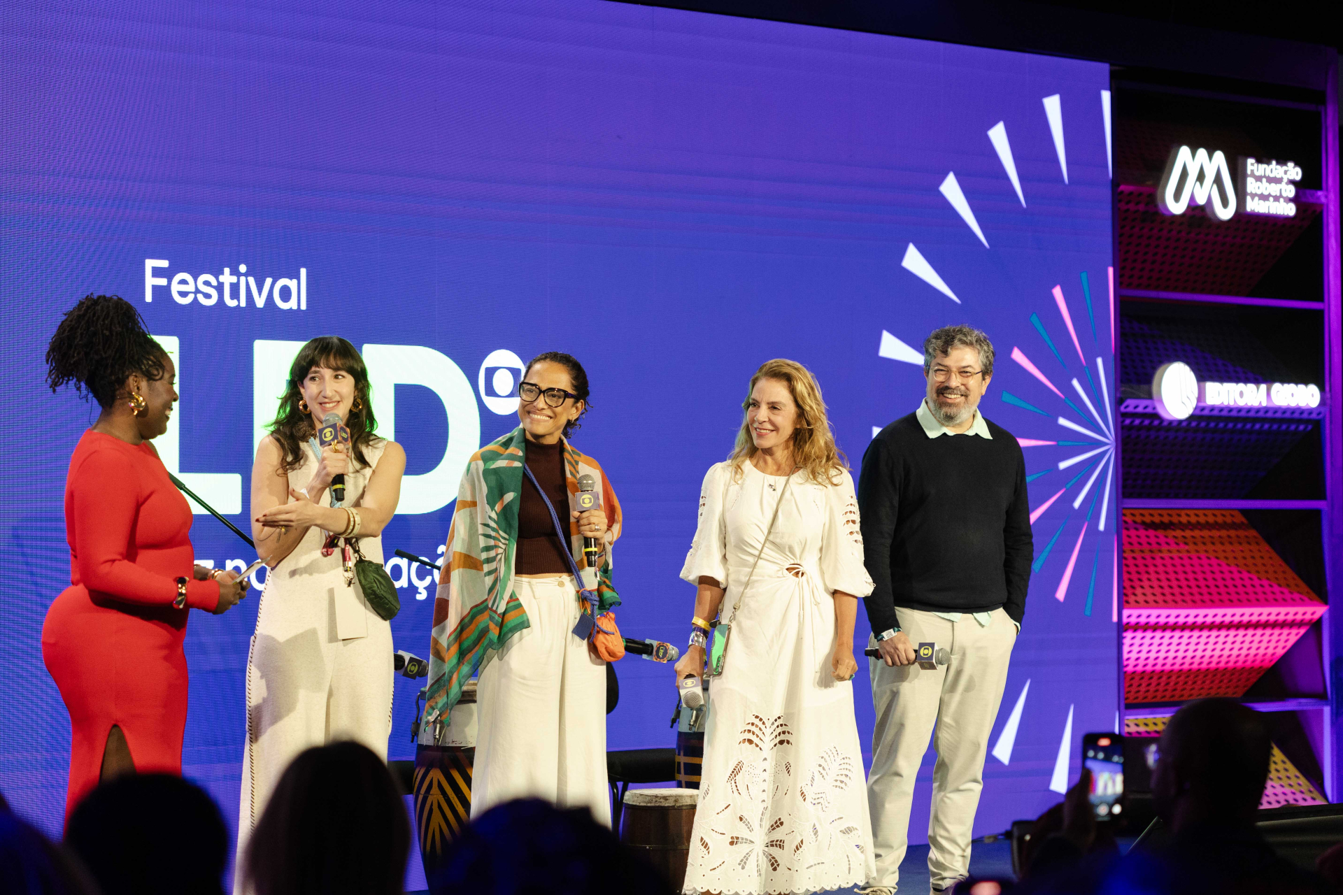 Cinco pessoas participam de uma conversa no palco do Festival LED, sorrindo e segurando microfones. Ao fundo, tela com o nome do festival e logotipos da Fundação Roberto Marinho e da Editora Globo. O grupo é composto por três mulheres e dois homens, todos em trajes casuais e elegantes, diante de uma plateia atenta.