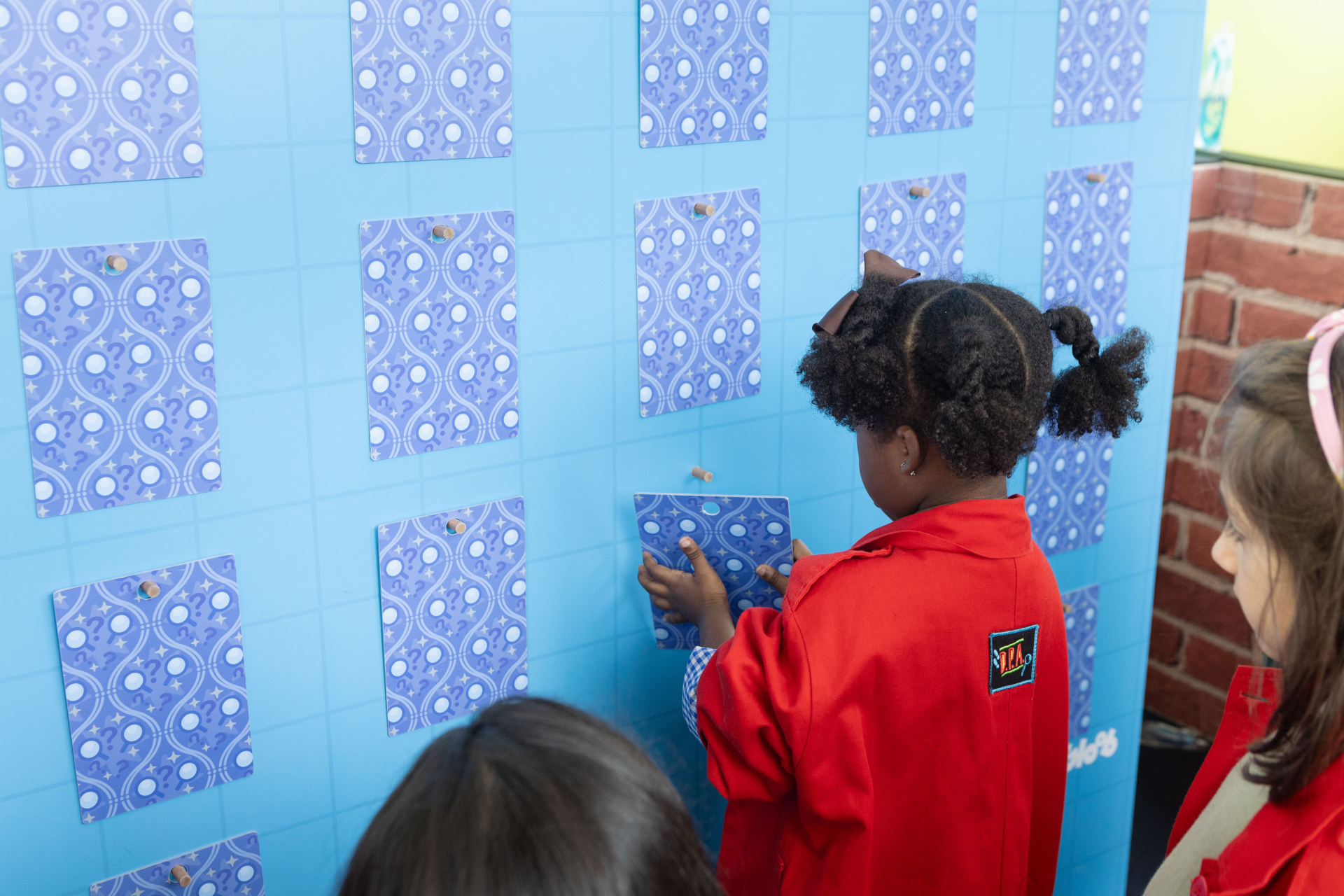 Duas meninas pequenas, usando jalecos vermelhos com o logo do DPA (Detetives do Prédio Azul) nas costas, participam de uma atividade interativa. Elas estão de frente para um grande painel azul com várias cartas viradas para baixo, decoradas com símbolos de interrogação. A menina ao centro toca uma das cartas, como se estivesse prestes a virá-la, enquanto a outra observa com curiosidade.