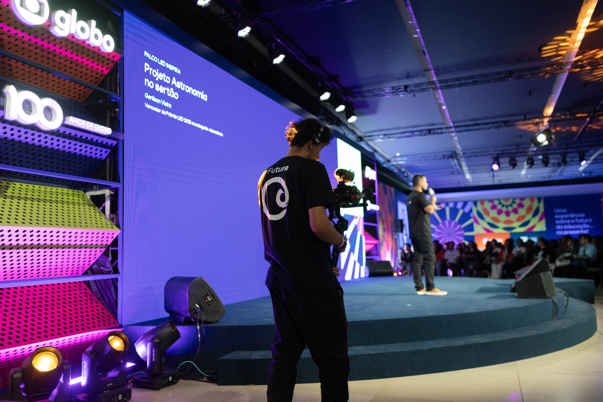 Ambiente interno de palco do Festival LED. Um homem faz uma apresentação no palco, enquanto um cinegrafista com camiseta do Canal Futura o filma de perto. No telão azul ao fundo, lê-se: “Palco LED Inspira – Projeto Astronomia no Sertão – Genilson Vieira – Vencedor do Prêmio LED 2023 na categoria educadores”.