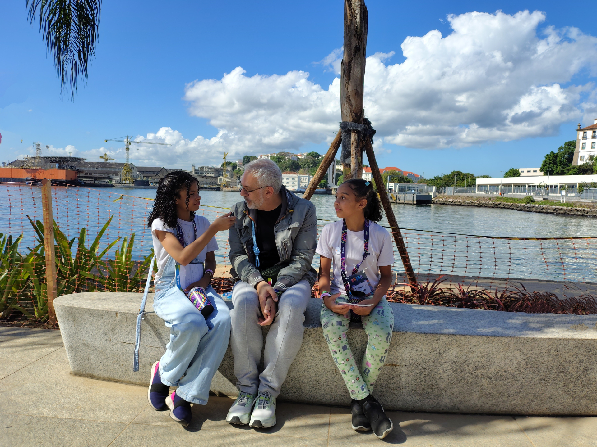 Três pessoas estão sentadas em um banco de pedra à beira de um rio, sob um céu azul com algumas nuvens brancas. À esquerda, uma menina de cabelos cacheados e escuros, vestindo uma camiseta branca e uma calça jeans clara larga, segura um pequeno microfone e entrevista o homem ao centro. Ela também usa tênis roxos e carrega uma bolsinha colorida. No meio, um homem de cabelo grisalho curto e barba branca, usando óculos escuros, uma jaqueta cinza e calça de moletom cinza, sorri e olha para a menina. À direita,