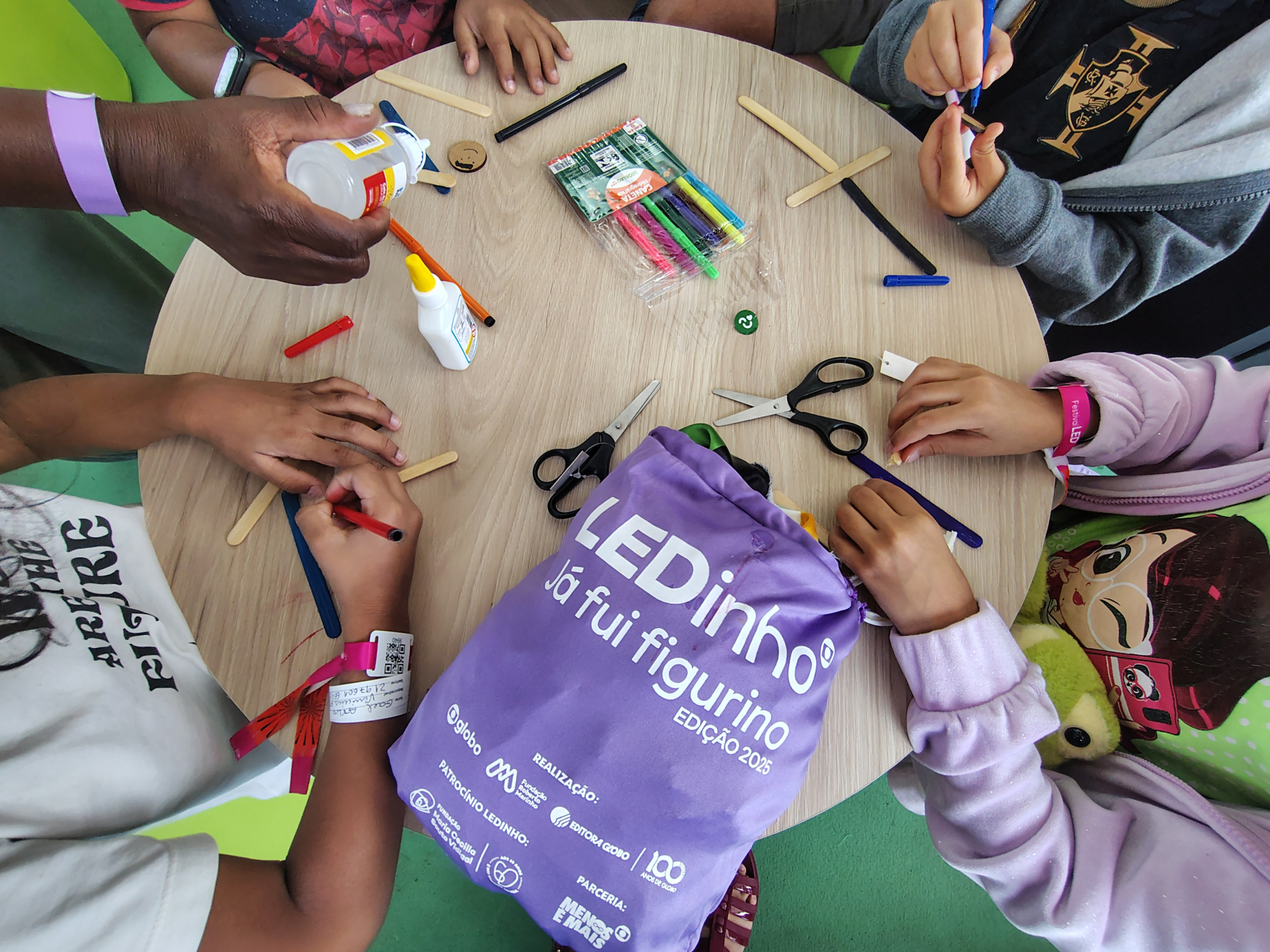Crianças participam de uma oficina de artesanato no Festival LEDinho, sentadas ao redor de uma mesa redonda. Elas utilizam materiais como palitos de madeira, tesouras, canetinhas e cola para criar suas produções. Ao centro da imagem, destaca-se uma mochila roxa com os dizeres “LEDinho – Já fui figurino – Edição 2025”. A cena transmite um clima de criatividade e aprendizado coletivo
