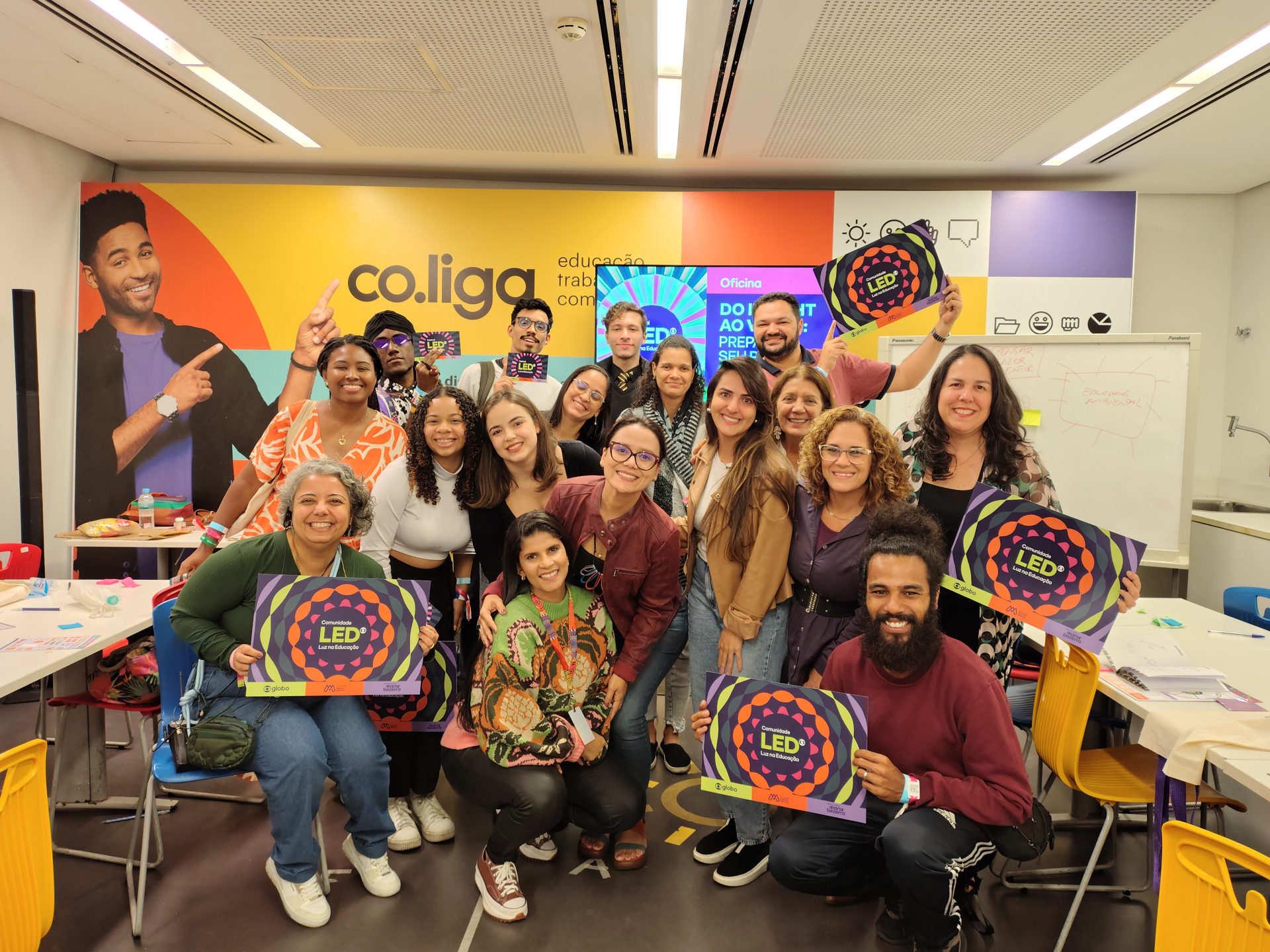 Grupo de cerca de 20 pessoas posa sorridente para a foto dentro de uma sala colorida com paredes decoradas com imagens e elementos gráficos. Ao fundo, há um painel com as palavras “co.liga” e uma televisão ligada. Algumas pessoas seguram cartazes do Festival LED – Luz na Educação. As mesas ao redor estão organizadas com papéis, marcadores e materiais de oficina. Todos parecem entusiasmados e engajados, transmitindo um clima de colaboração e celebração.