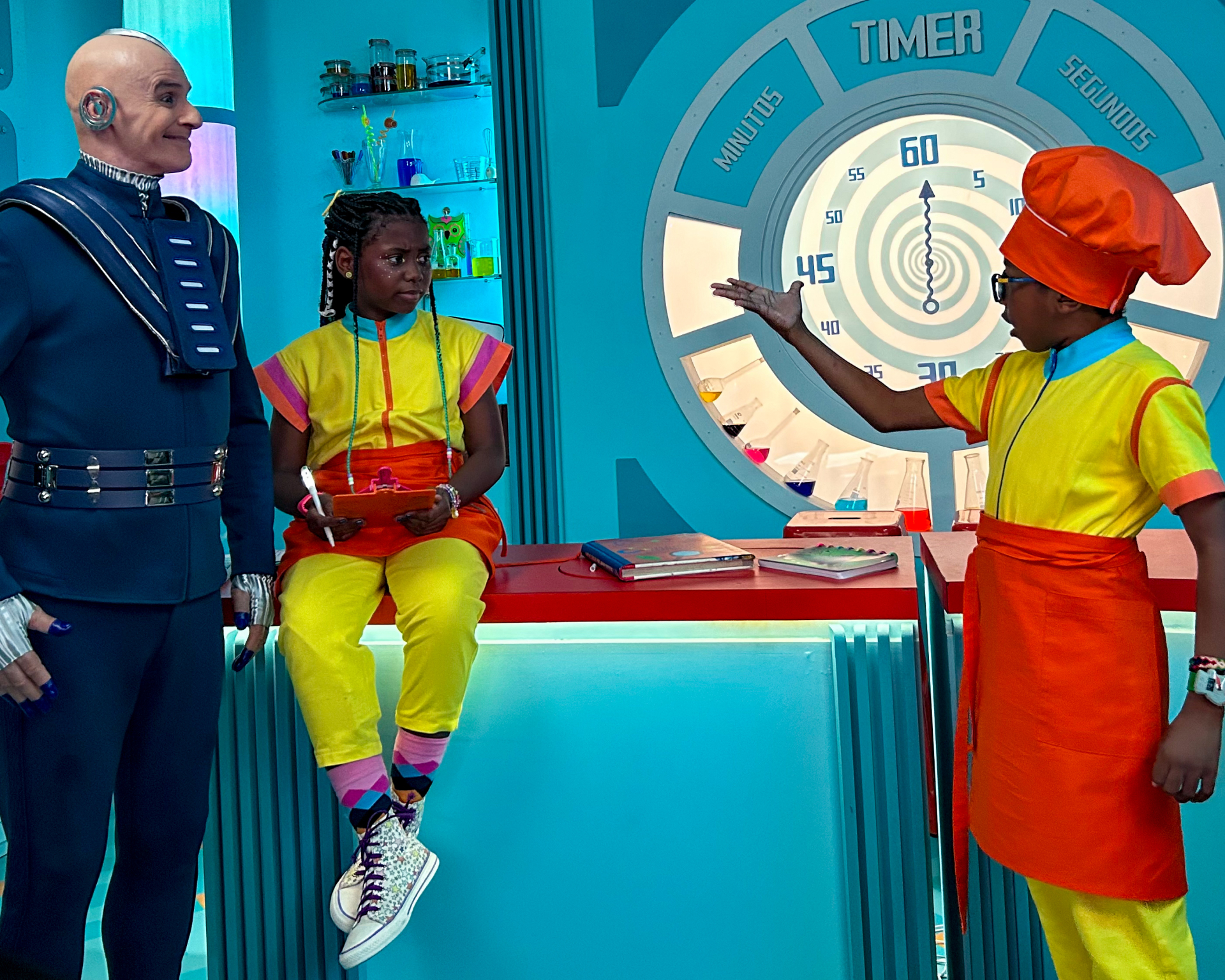 Três pessoas estão em um ambiente futurista e colorido. Duas crianças negras, uma menina e um menino, usam macacões amarelos com detalhes coloridos, e estão de frente para um homem careca vestido com uma roupa azul escura e futurista. A menina está sentada em um balcão vermelho com um tablet nas mãos, enquanto o menino, usando um chapéu de cozinheiro laranja, gesticula com uma das mãos, como se estivesse explicando algo. Atrás deles, há um grande cronômetro azul com ponteiros marcando os segundos e vários f