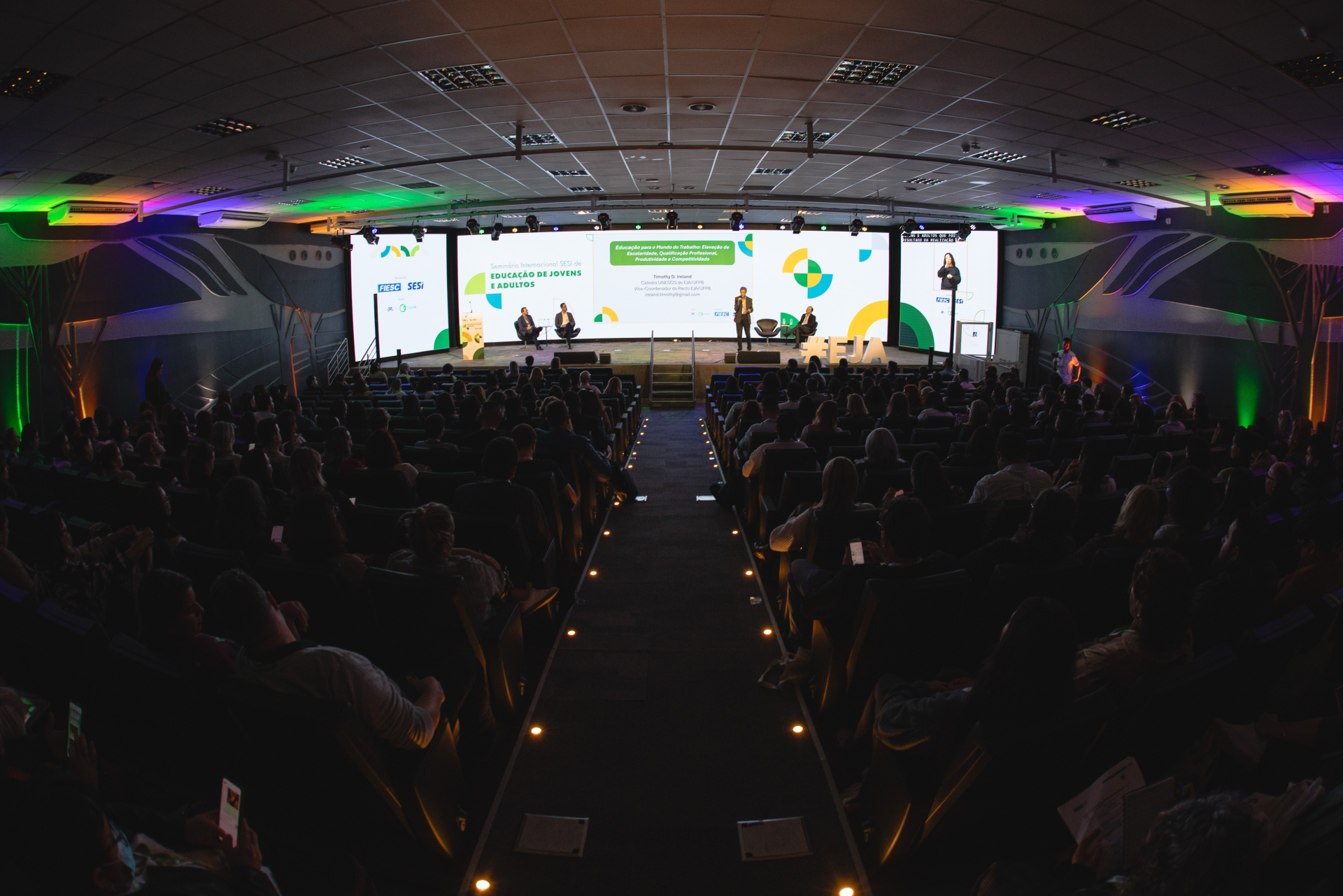 Auditório escuro e cheio de pessoas sentadas, assistindo a uma apresentação no palco iluminado. À frente, cinco pessoas participam de um painel no “Seminário Internacional SESI de Educação de Jovens e Adultos”. Um grande telão exibe o tema “Educação para a inclusão no trabalho: formação de pessoas jovens e adultas com deficiência”. À direita, uma intérprete de Libras aparece em tela vertical. O ambiente tem iluminação decorativa nas cores verde, roxa e laranja, criando uma atmosfera moderna. No palco, letra