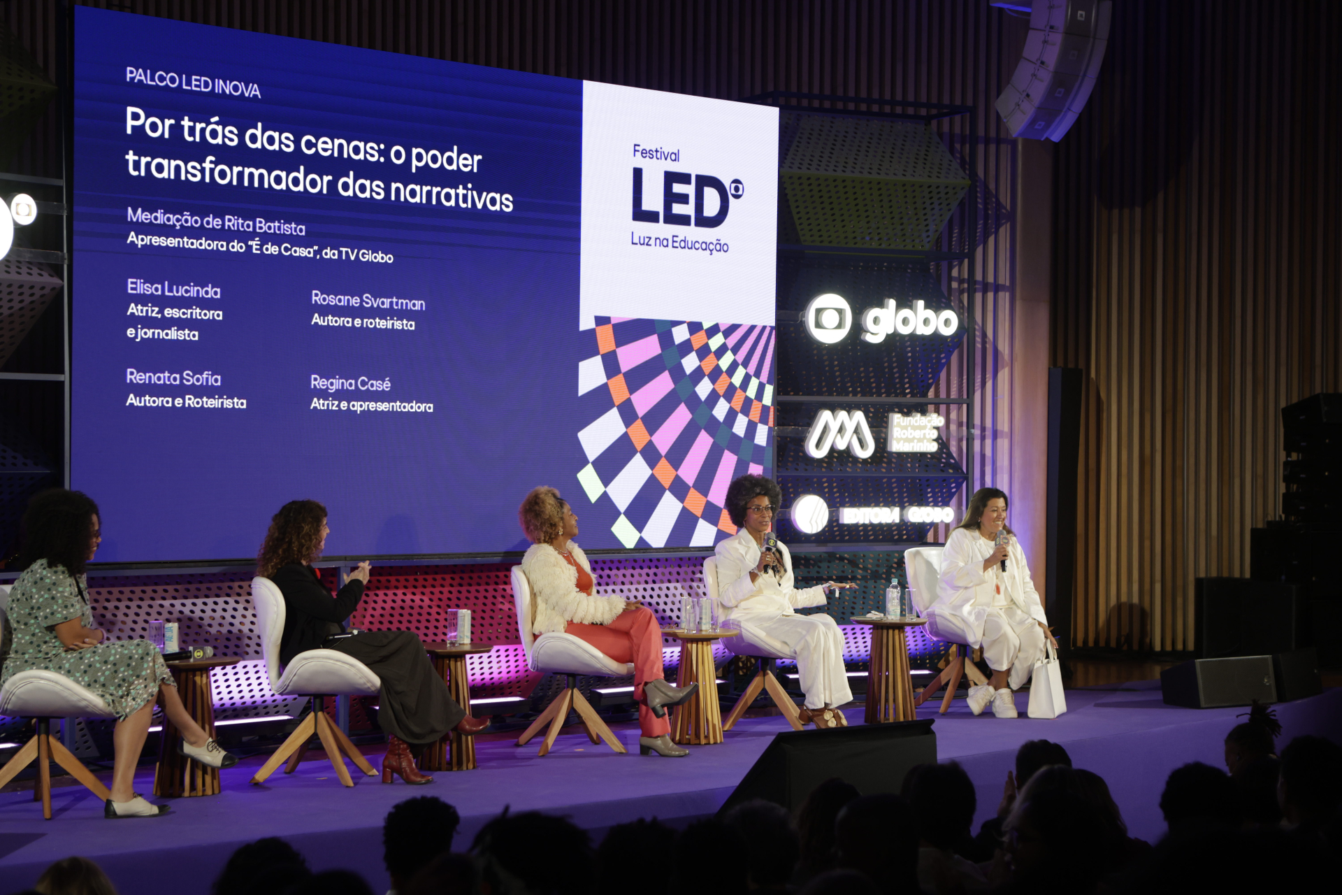 Painel “Por trás das cenas: o poder transformador das narrativas”, realizado no Palco LED Inova durante o Festival LED – Luz na Educação. Seis mulheres estão sentadas em poltronas no palco roxo, participando da conversa. À esquerda, um grande telão exibe os nomes das participantes: Rita Batista (mediadora), Elisa Lucinda, Rosane Svartman, Renata Sofia, Regina Casé. Todas seguram microfones, envolvidas na discussão. Ao fundo, logotipos da Globo, Fundação Roberto Marinho e Editora Globo estão iluminados. A pl