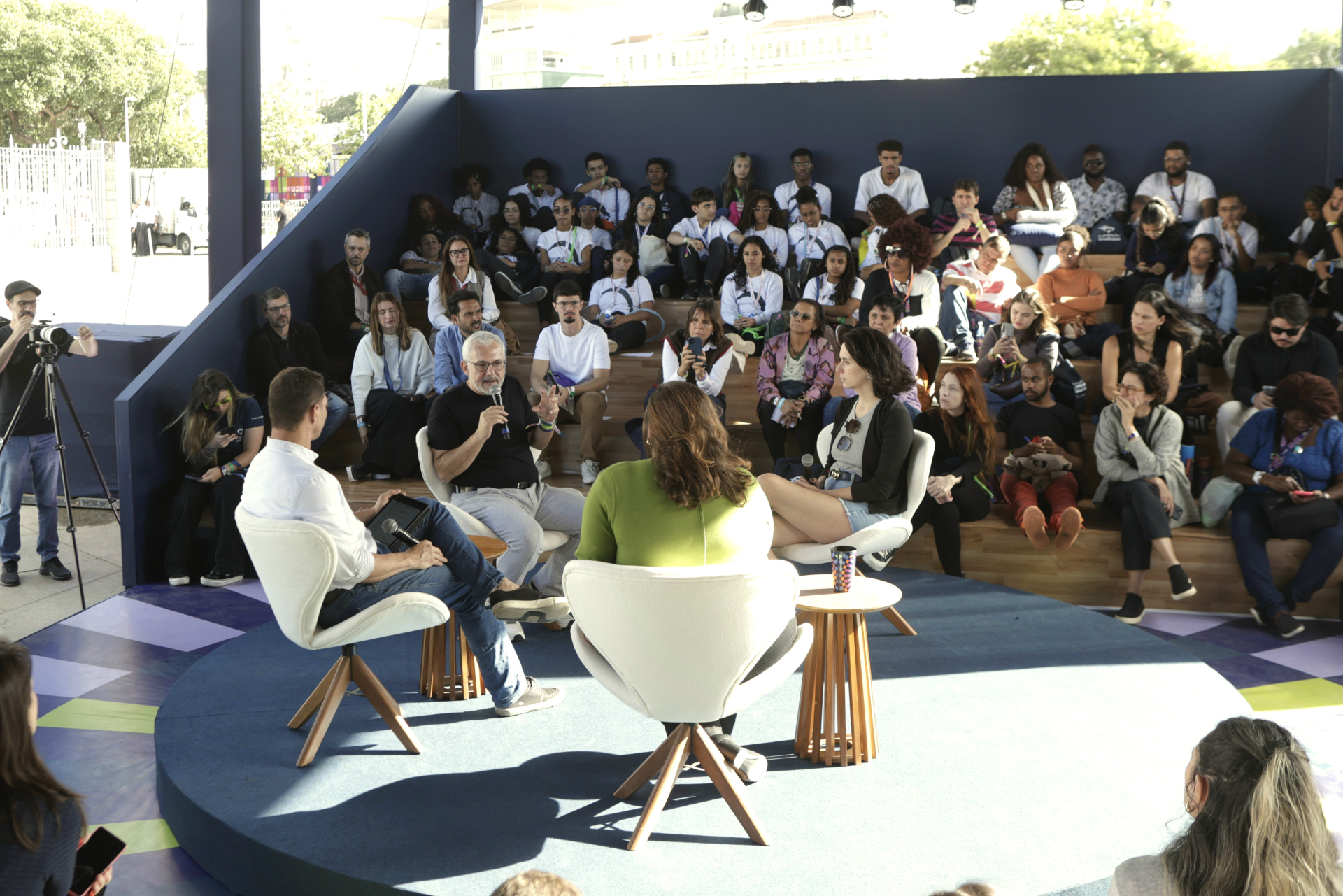 Espaço de conversa no Festival LED – Luz na Educação, com plateia disposta em arquibancada de madeira. Quatro pessoas participam de um bate-papo no centro, sentadas em poltronas brancas sobre palco circular azul. Uma delas, um homem de cabelos grisalhos e barba, fala ao microfone enquanto gesticula. Ao redor, dezenas de pessoas assistem com atenção, incluindo jovens estudantes, adultos e profissionais da área de educação. A estrutura é coberta e iluminada pela luz natural, criando um ambiente acolhedor e pa