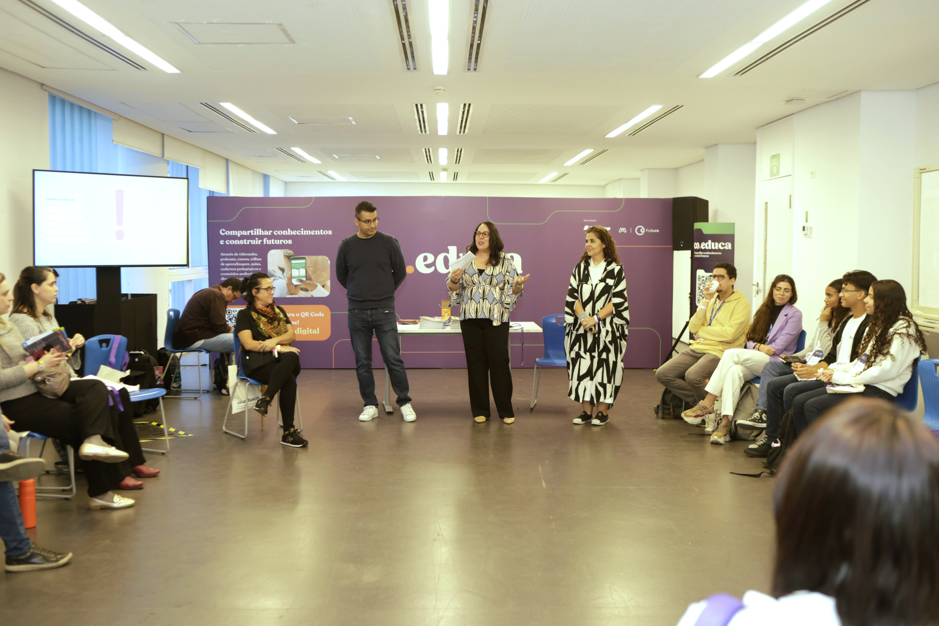 Cerca de 20 pessoas participam de uma roda de conversa em uma sala ampla, com iluminação natural e paredes brancas. Três pessoas — duas mulheres e um homem — estão em pé ao centro, conduzindo a atividade. À direita e à esquerda, os participantes estão sentados em cadeiras azuis, prestando atenção ou anotando. Ao fundo, um painel roxo exibe a frase “Compartilhar conhecimentos e construir futuros” ao lado da marca “co.educa”. Um telão e banners também fazem parte da ambientação. O clima é de escuta ativa, tro