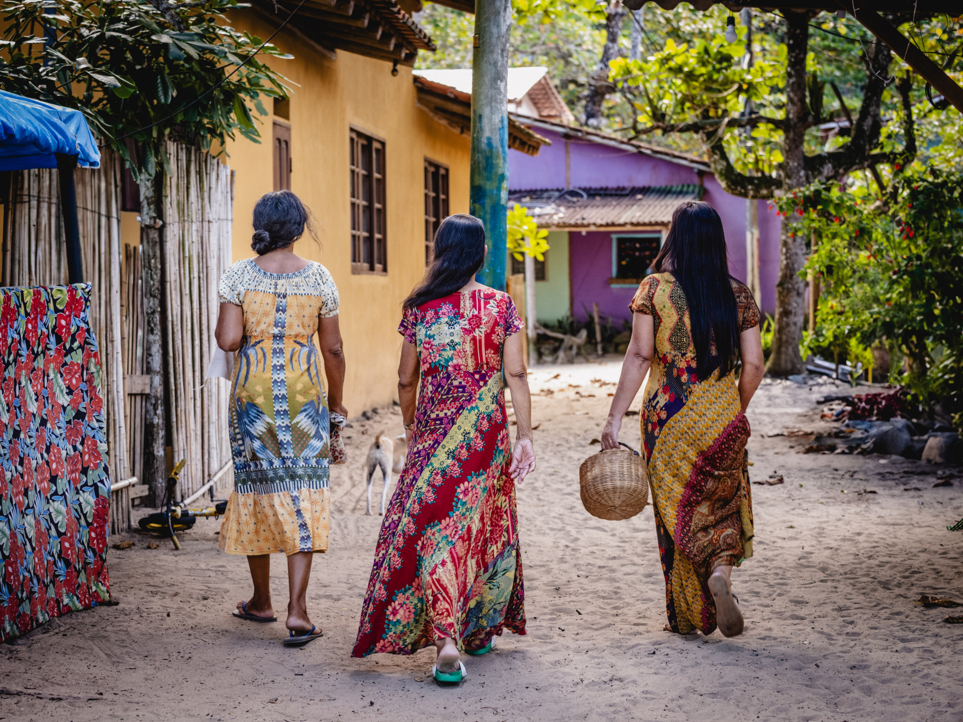 Três mulheres caminham de costas por uma rua de terra em uma vila, cercadas por árvores e casas coloridas. Elas usam vestidos longos e estampados com cores vibrantes. Uma delas segura uma cesta de palha, e um cachorro aparece ao fundo, entre elas. As casas têm cores vivas, como amarelo e lilás, com janelas e portas de madeira. A cena transmite tranquilidade e conexão com a cultura local.