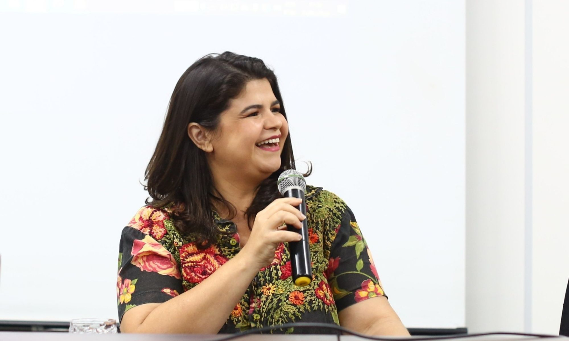 Uma mulher de cabelos escuros e lisos, com uma camisa estampada floral colorida, sorri amplamente enquanto segura um microfone. Ela está sentada em uma mesa de debate, olhando para o lado direito da imagem. O fundo é claro, com projeção de tela branca.
