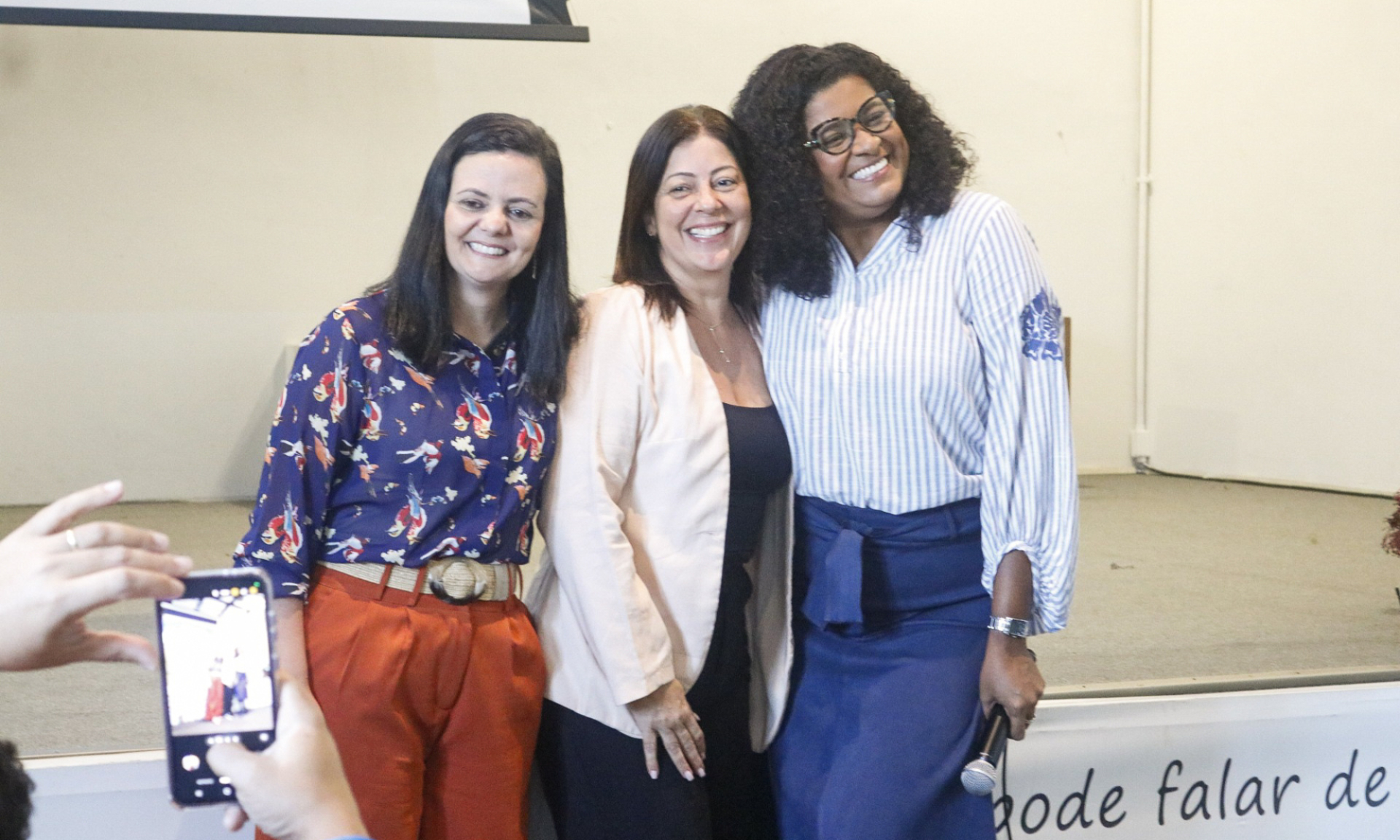 Três mulheres posam sorrindo em frente a um palco. A da esquerda veste blusa estampada azul-marinho com flores vermelhas e calça vermelha; a do centro usa blazer claro e blusa preta; a da direita, blusa listrada azul e branca com um detalhe floral na manga, e uma saia azul. Ela segura um microfone. À frente, uma pessoa fotografa o trio com um celular. O fundo é claro, com parte de uma frase visível na parede: “pode falar de...”. A cena sugere encerramento ou homenagem após uma fala ou apresentação.