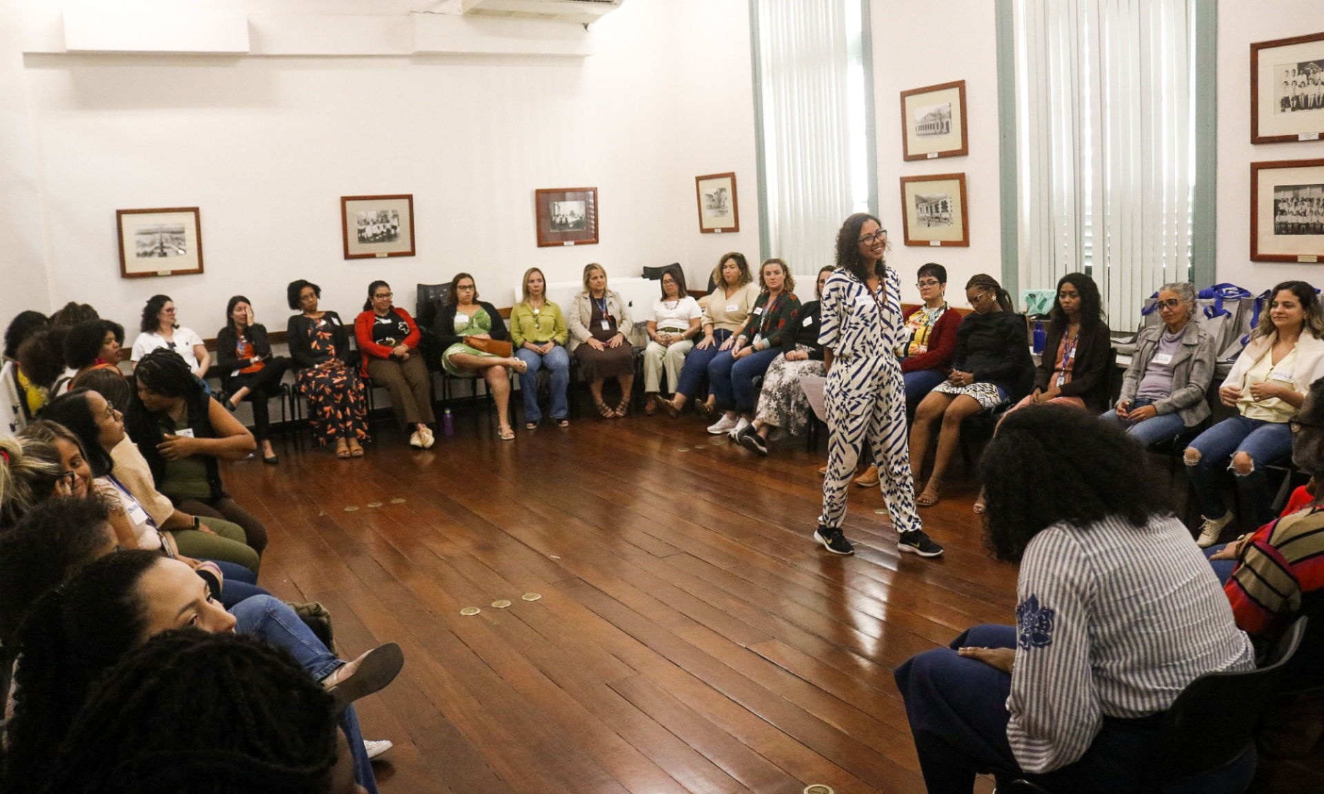 Sala com piso de madeira e paredes brancas decoradas com quadros emoldurados em tom escuro. Cerca de 30 pessoas estão sentadas formando um círculo, olhando para uma mulher que está em pé no centro, falando e sorrindo. Ela usa um macacão estampado em azul e branco. As pessoas ao redor são majoritariamente mulheres, com expressões atentas, demonstrando escuta ativa. O ambiente sugere um momento de partilha ou roda de conversa.