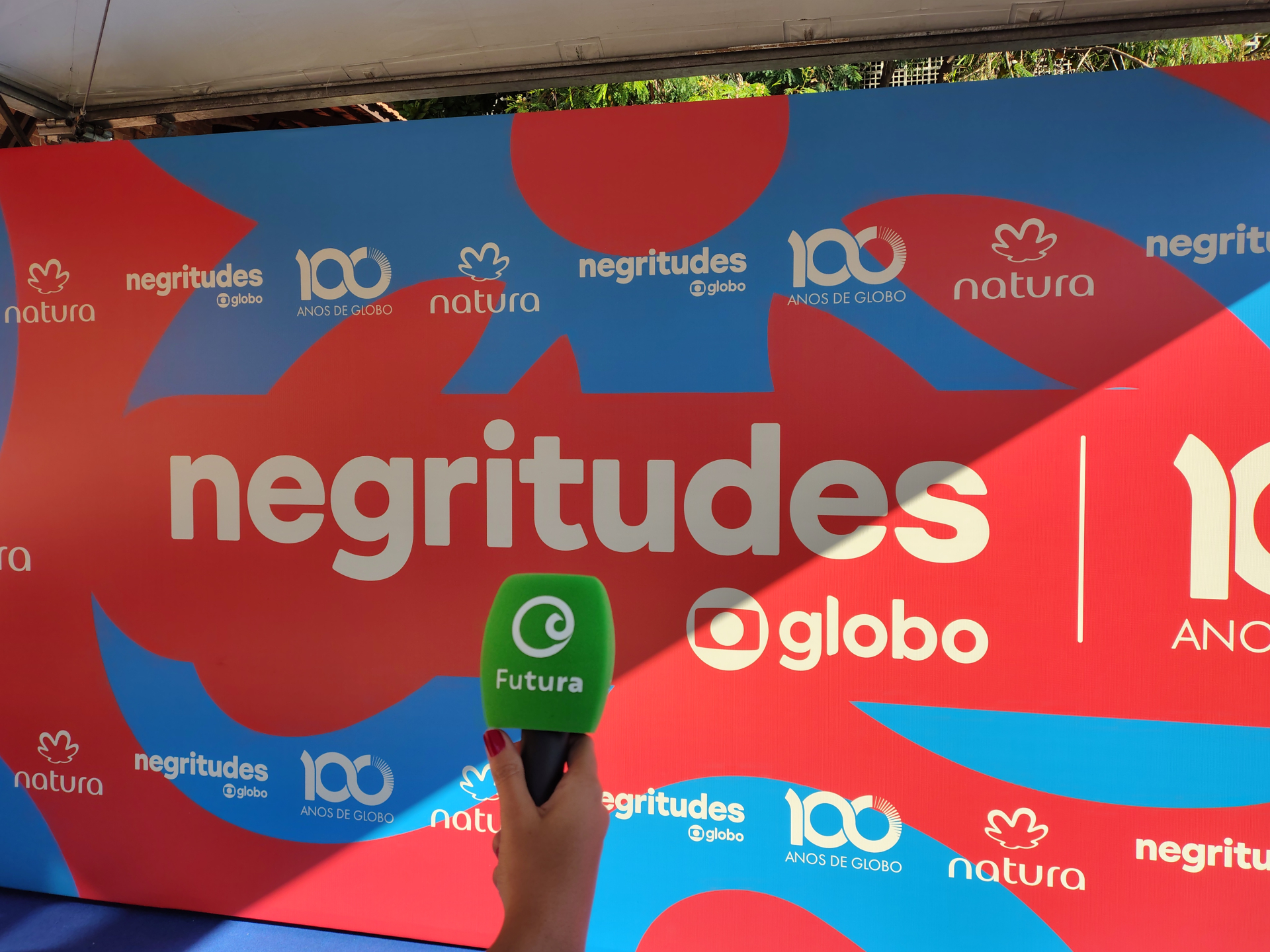 Painel colorido com fundo em azul e vermelho, estampado com logomarcas e palavras relacionadas ao evento. No centro, em letras grandes e brancas, está escrito “negritudes” com o logotipo da Globo abaixo. Ao redor, repetem-se as logomarcas da Natura (uma folha estilizada), “100 anos de Globo” e o nome “negritudes @globo”. Em primeiro plano, na parte inferior central da imagem, uma mão segura um microfone com espuma verde, onde está escrito “Futura” em branco, com o logotipo do Canal Futura. A imagem foi tira
