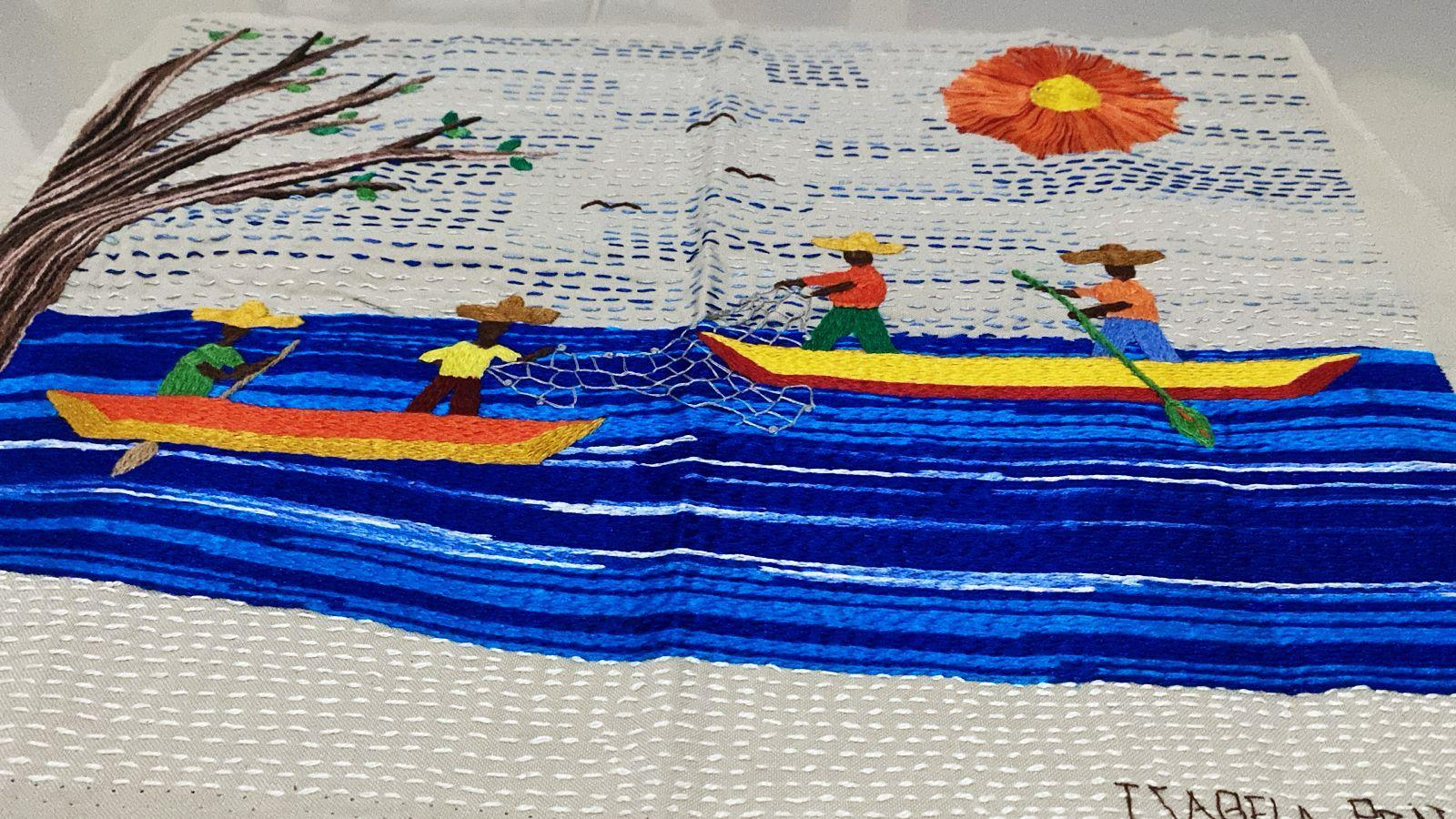Bordado vibrante em tecido claro mostra duas canoas coloridas flutuando sobre ondas azuis. Em uma delas, duas pessoas pescam com rede, e na outra, duas pessoas remam. Ao fundo, um grande sol laranja com centro amarelo brilha no céu. Um galho de árvore aparece à esquerda e pequenos pássaros pretos voam ao fundo. A cena representa a pesca artesanal em um dia ensolarado.