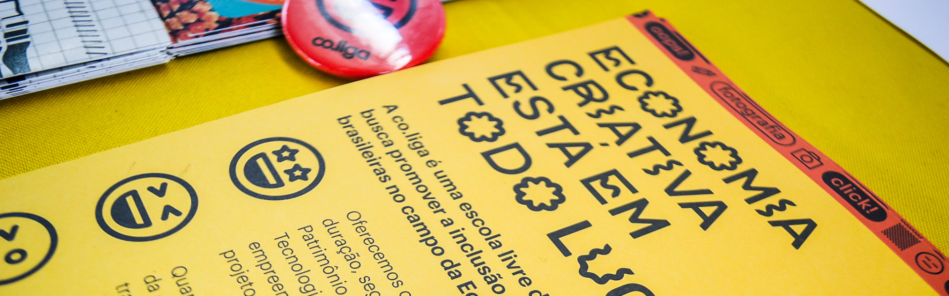 Material gráfico promocional sobre economia criativa disposto sobre uma superfície amarela. Em destaque, um folder amarelo com o título "ECONOMIA CRIATIVA ESTÁ EM TODO LUGAR" escrito com letras estilizadas. Abaixo, há um texto explicativo sobre a iniciativa Co.liga, uma escola livre digital gratuita voltada à inclusão produtiva de juventudes no campo da economia criativa. Em cima do folder, estão bottons coloridos, sendo o principal um azul com ícone de câmera e o nome "Co.liga", indicando uma abordagem vol