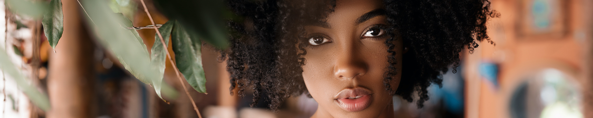 Uma linda e jovem mulher negra de cabelos cacheados no estilo black power. Ela está bem maquiada, olhando diretamente para a câmera e tocando uma plantinha com a mão direita