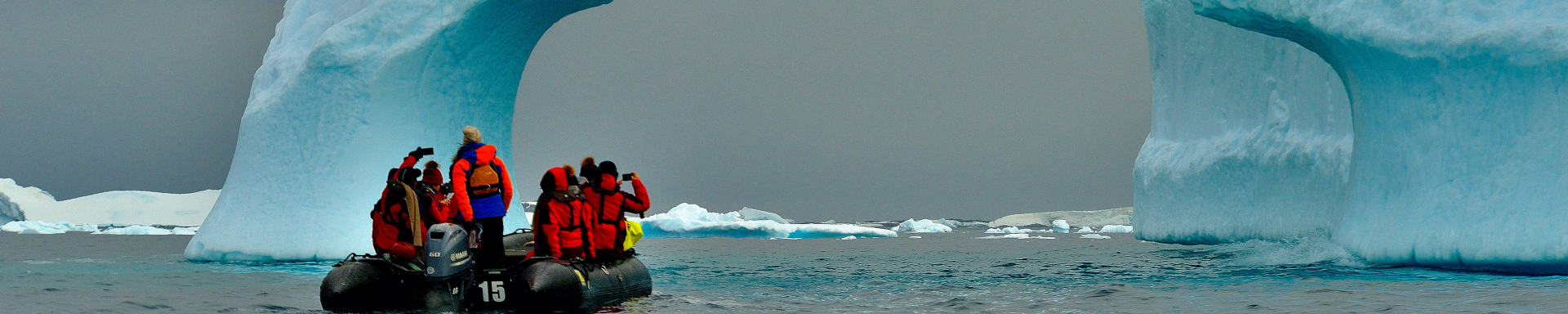 Um bote inflável com algumas pessoas muito agasalhadas está navegando em um mar gélido da Antártida e está se aproximando de uma espécie de iceberg com uma abertura