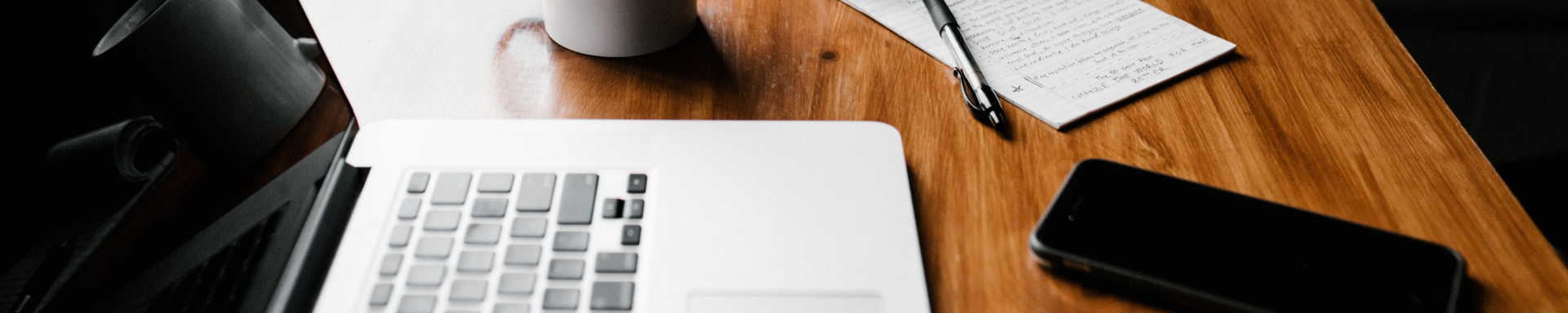 Um notebook aberto e desligado, uma xícara branca de café, um bloco de anotações, uma caneta e um celular estão em cima de uma mesa de madeira sendo iluminada pela luz solar que vem da janela ao lado