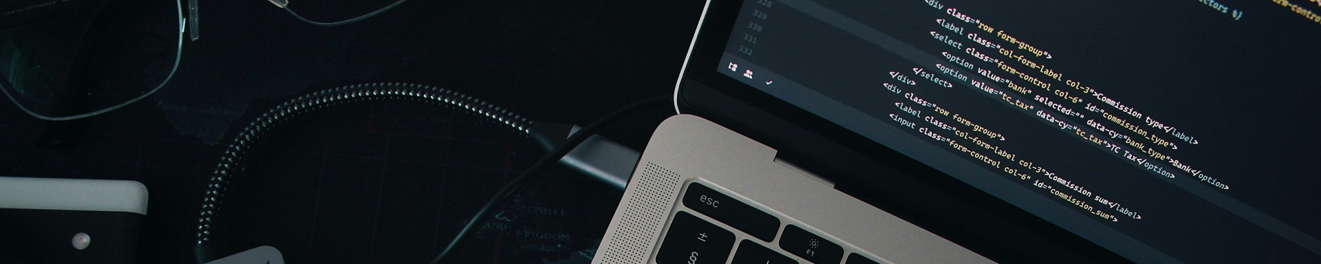 Um notebook visto parcialmente, aberto e exibindo uma tela com código de programação. O teclado preto com teclas iluminadas está em destaque. Ao lado do notebook, há um adaptador USB-C cinza conectado por um cabo. Também é possível ver um par de óculos, um caderno branco e um mapa-múndi estilizado como superfície da mesa. A iluminação é suave, sugerindo um ambiente de trabalho moderno e tecnológico.