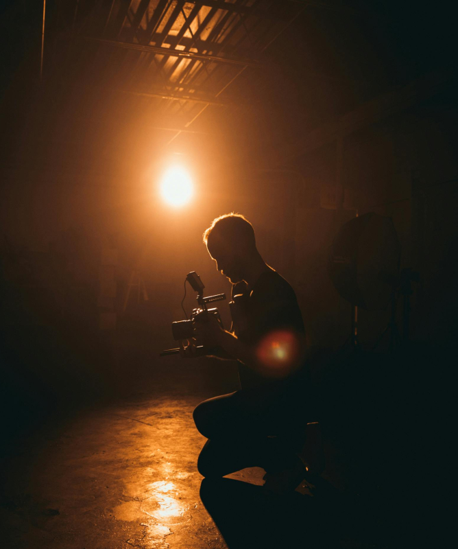 Em um ambiente escuro e ligeiramente esfumaçado (mais parece uma neblina, talvez), iluminado por uma lâmpada ao fundo, vê-se a silhueta de um homem agachadinho, segurando uma câmera.