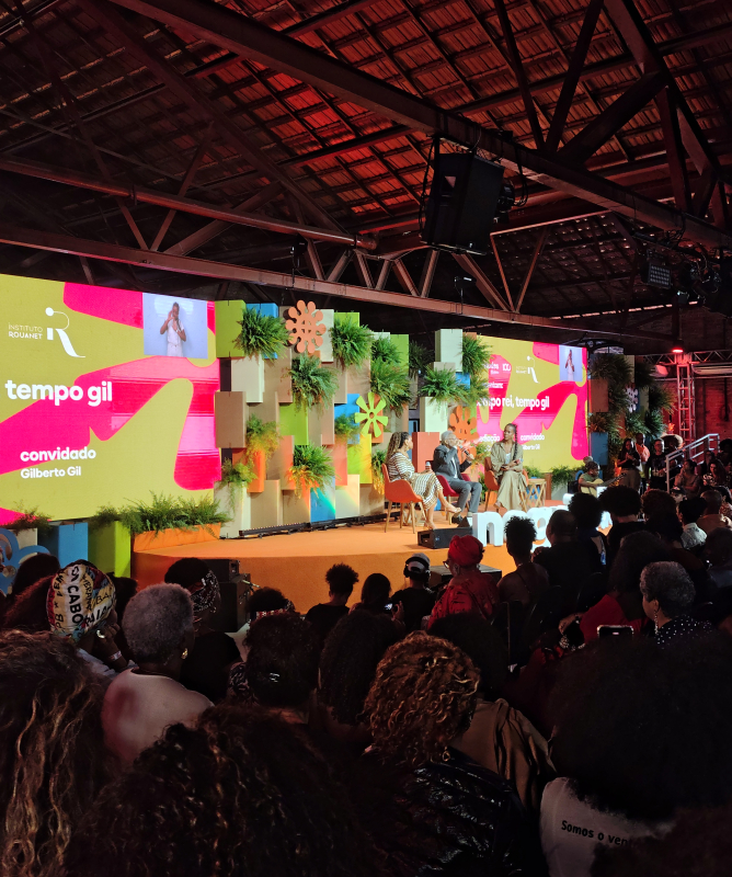 Auditório lotado assiste ao painel "Tempo Rei, Tempo Gil", durante o evento Negritudes Globo. No palco, decorado com plantas e painéis coloridos, estão sentados Gilberto Gil, a jornalista Maju Coutinho e a cantora Teresa Cristina, que conduzem a conversa. Um telão ao fundo exibe o nome do painel, os nomes dos participantes e as marcas dos organizadores. O ambiente é coberto, com estrutura de madeira e iluminação quente. 