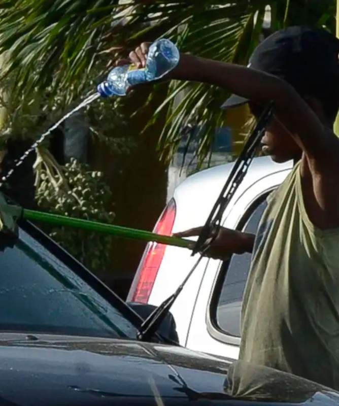 Um menino negro, magro e com boné escuro está limpando o para-brisa de um carro com uma esponja presa a um cabo verde, enquanto derrama água de uma garrafa plástica. Ele aparenta estar trabalhando na rua, sob sol, cercado por carros e vegetação ao fundo.