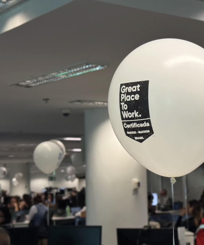 Ambiente interno de escritório com diversas pessoas concentradas em seus computadores, trabalhando em um espaço moderno, iluminado e com layout aberto. No centro da imagem, um balão branco com a frase “Great Place To Work – Certificada – Mai/2023 - Mai/2025 – BRASIL” em destaque, indicando o reconhecimento da empresa como um excelente local para trabalhar. Ao fundo, vê-se o logo estilizado da instituição em grande escala na parede e funcionários ocupando diferentes estações de trabalho. O clima é de rotina 
