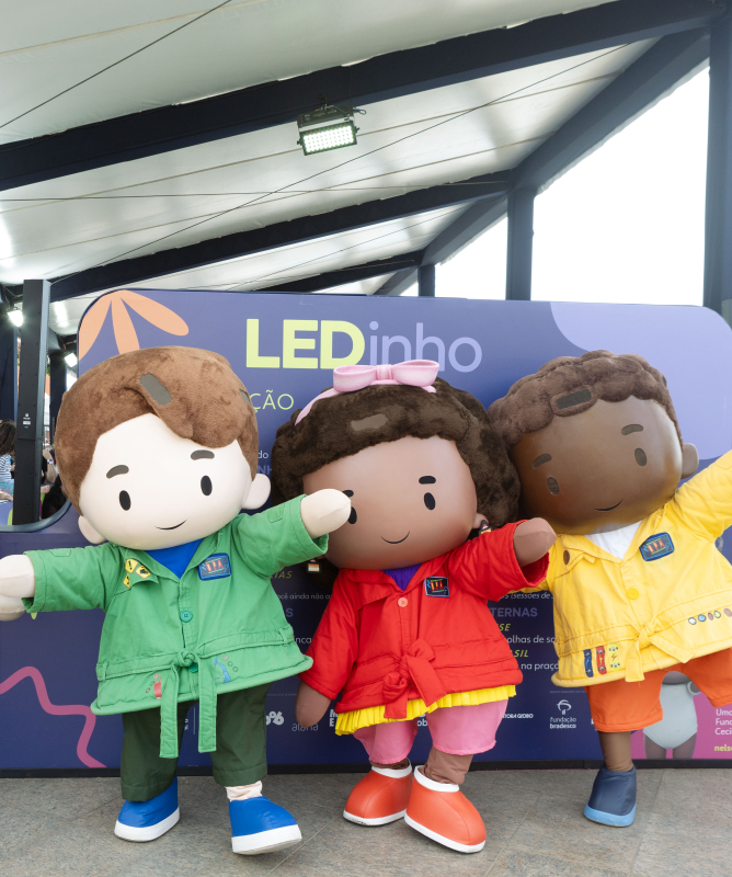 Quatro personagens infantis em tamanho gigante posam sorridentes, de mãos dadas, em frente a um painel com a identidade visual do LEDinho, espaço infantil do Festival LED. Os mascotes usam jalecos coloridos (verde, vermelho, amarelo e um vestido roxo com estampa de planetas) e representam crianças diversas. Ao fundo, há árvores, uma tenda coberta e uma bancada de atendimento com pessoas circulando.