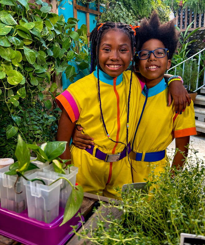 Duas crianças estão abraçadas e sorrindo em um jardim cheio de plantas verdes. Estão vestindo os mesmos macacões amarelos com detalhes coloridos. Atrás delas há uma escada de azulejos e muitas plantas ao redor. Em frente, sobre uma bancada de madeira, há vasos e mudas com etiquetas, indicando um espaço de cultivo e aprendizado sobre plantas.