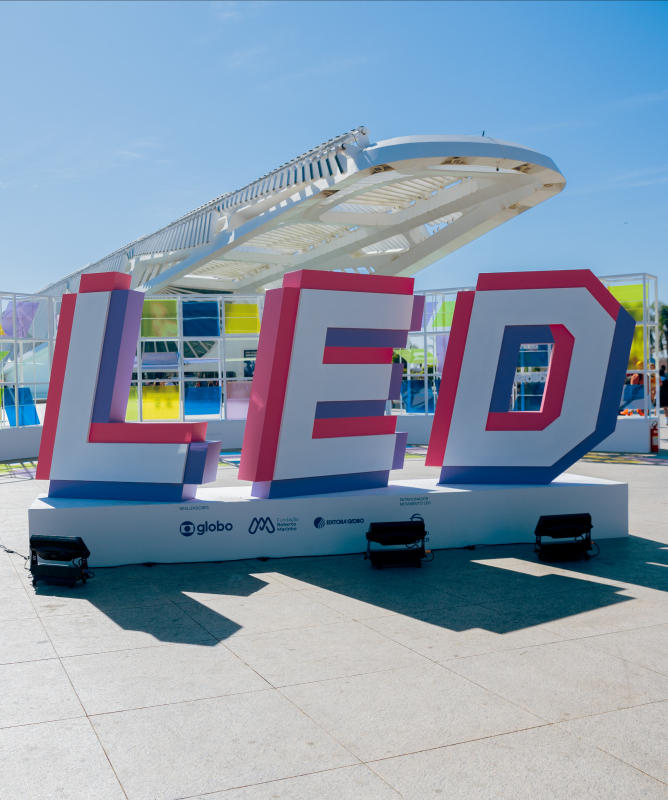 Em uma ampla área externa sob céu azul e ensolarado, destaca-se uma grande instalação com as letras tridimensionais “LED” em cores vibrantes (rosa, branco, roxo e azul), posicionada diante do Museu do Amanhã, no Rio de Janeiro. Ao fundo, uma estrutura colorida com painéis translúcidos em tons de amarelo, verde, azul e roxo exibe os logotipos da Globo, Fundação Roberto Marinho e Editora Globo, realizadores do evento.