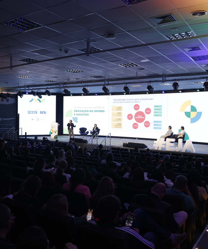 Auditório moderno com cadeiras ocupadas por diversas pessoas assistindo a uma apresentação. No palco iluminado, há quatro pessoas — duas em pé e duas sentadas — participando de um painel com telão ao fundo. No telão está escrito: “Seminário Internacional SESI de Educação de Jovens e Adultos”, com gráficos e textos explicativos. À direita, uma tela vertical mostra uma intérprete de Libras (Língua Brasileira de Sinais). O ambiente tem iluminação colorida nas laterais e teto com painéis de LED.