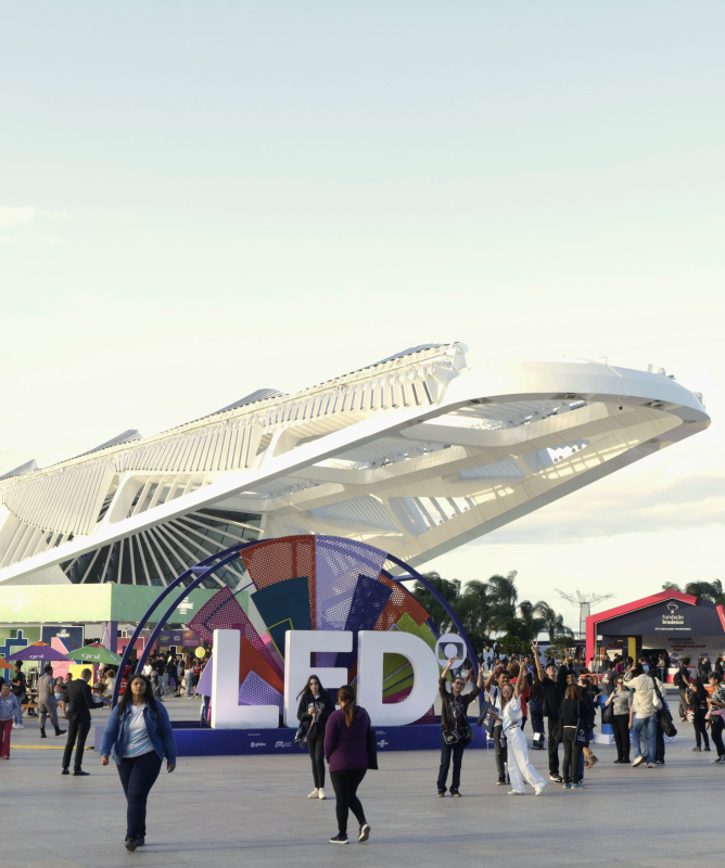 Vista ampla da área externa do Museu do Amanhã, no Rio de Janeiro, durante o Festival LED – Luz na Educação. Em primeiro plano, pessoas caminham e interagem ao redor de uma grande instalação colorida com as letras “LED”. Ao fundo, o mar, palmeiras e tendas coloridas compõem o cenário festivo.