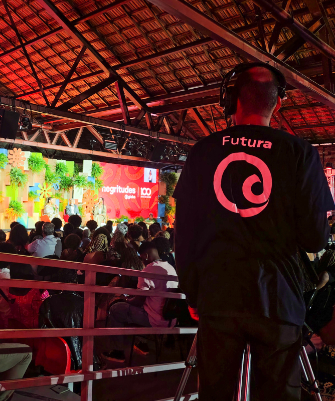 A imagem mostra um auditório coberto, com teto de madeira e vigas aparentes, lotado de pessoas assistindo a um evento. À frente do público, há um grande palco decorado com painéis coloridos e plantas verdes. Um telão vermelho exibe as palavras “negritudes @globo” e “100 anos de Globo” em branco. Há também pequenas telas que mostram a imagem de uma apresentadora.  À direita da imagem, em primeiro plano, um cinegrafista do Canal Futura está de costas, operando uma câmera sobre um tripé. Ele veste uma camisa p