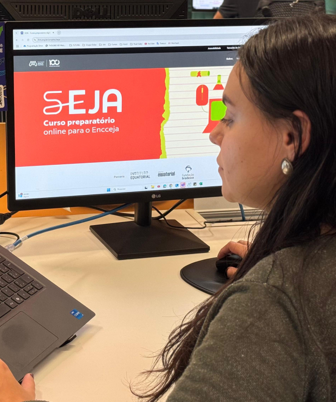 Uma mulher de cabelos longos e escuros, usando brinco de prata, está sentada em frente a duas telas em um ambiente de escritório. Ela olha para um monitor onde está aberto o site do curso “SEJA – Curso preparatório online para o Encceja”. À sua esquerda, há um notebook com a página inicial da Plataforma Futura aberta, exibindo programas como Co.liga, Telê Curso e Aprendê Legal. No fundo, outras pessoas também estão estudando. O ambiente é moderno, com iluminação clara e clima de concentração.