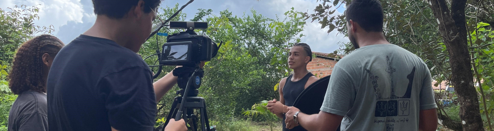 Equipe de filmagem trabalha em um ambiente com árvores e chão de terra.